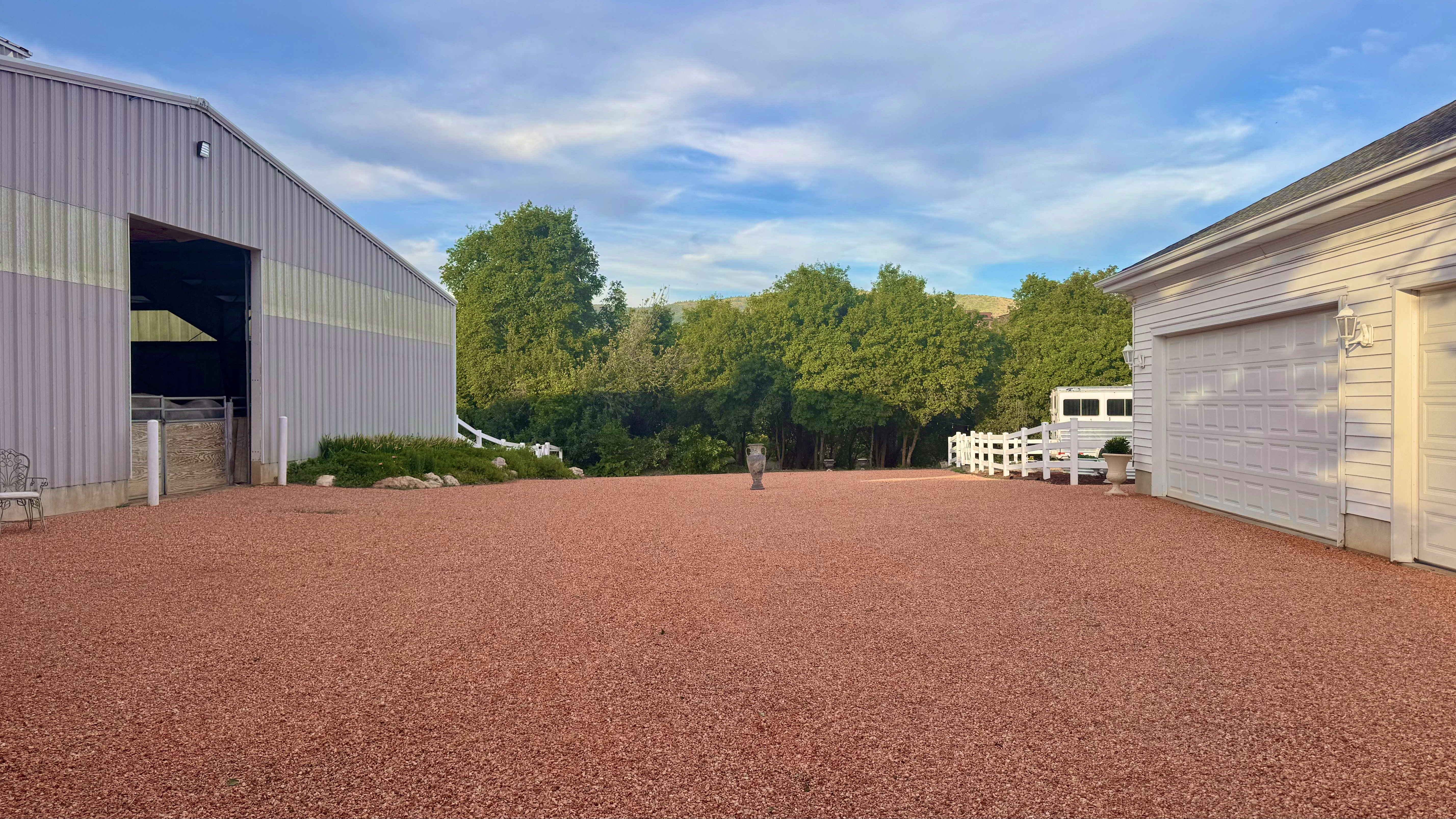 Expansive red gravel area with a large metal-sided outbuilding featuring an open entryway and a white-sided two-car garage with overhead doors