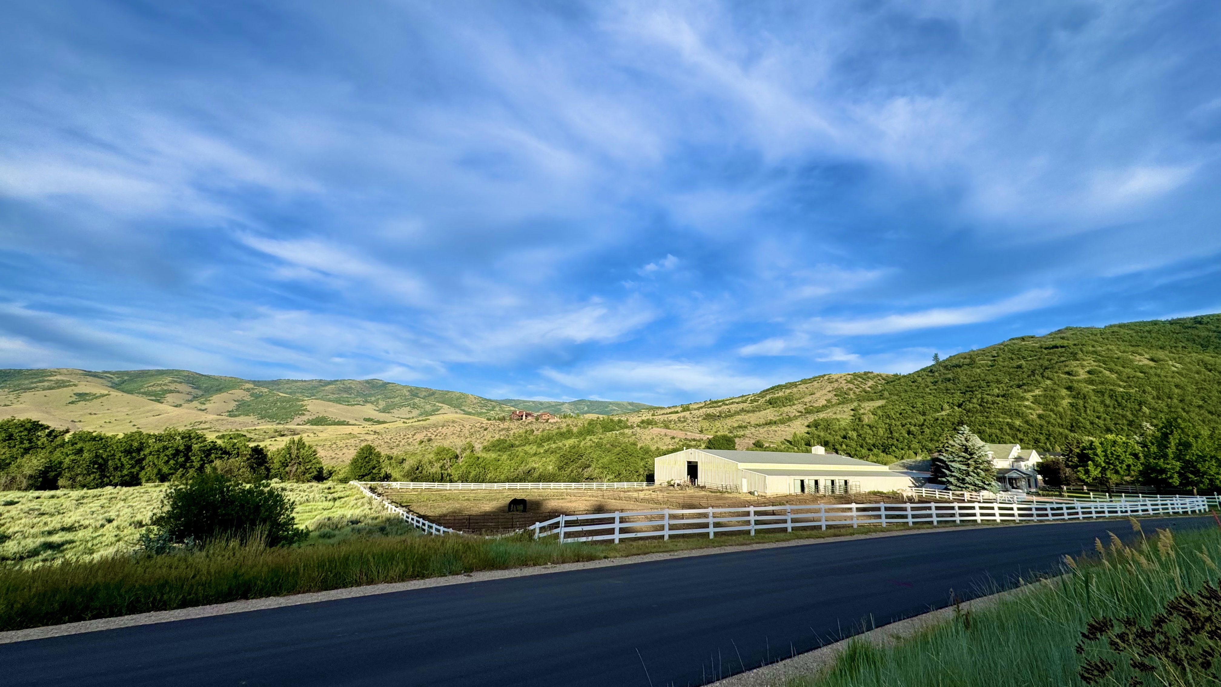Expansive rural property featuring a large barn structure, a white-fenced perimeter, and a residential building with a prominent evergreen tree