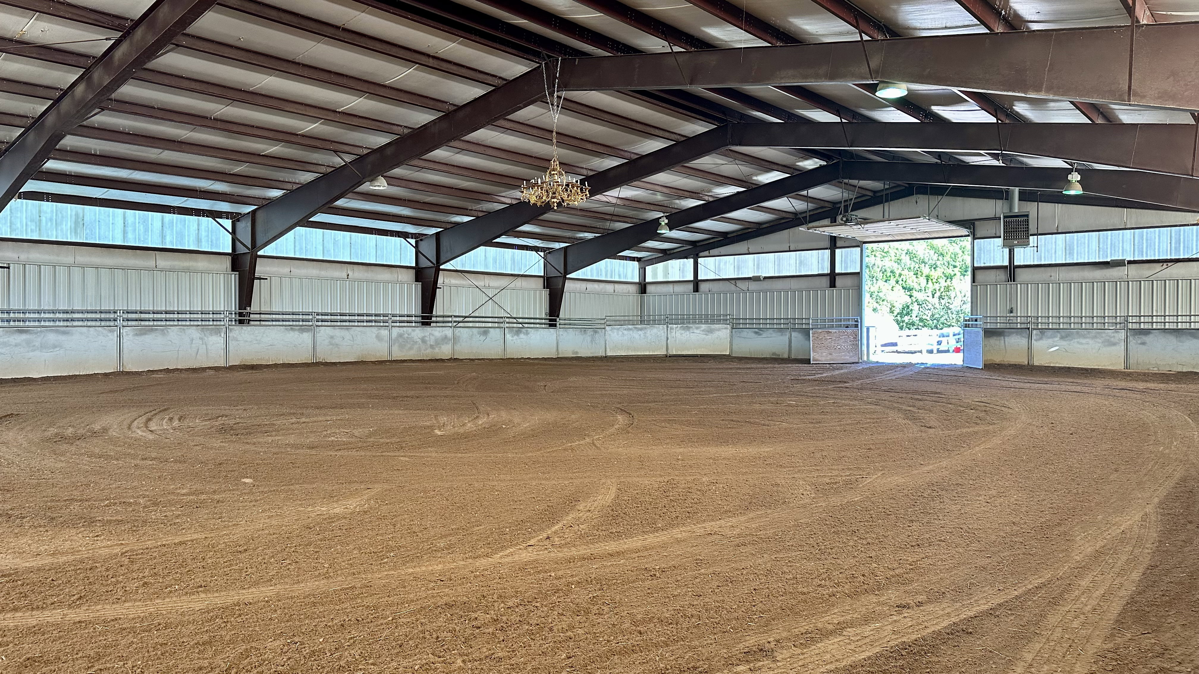 Expansive indoor arena featuring a sand surface, high ceilings with exposed steel beams, perimeter railing, and a large entry door