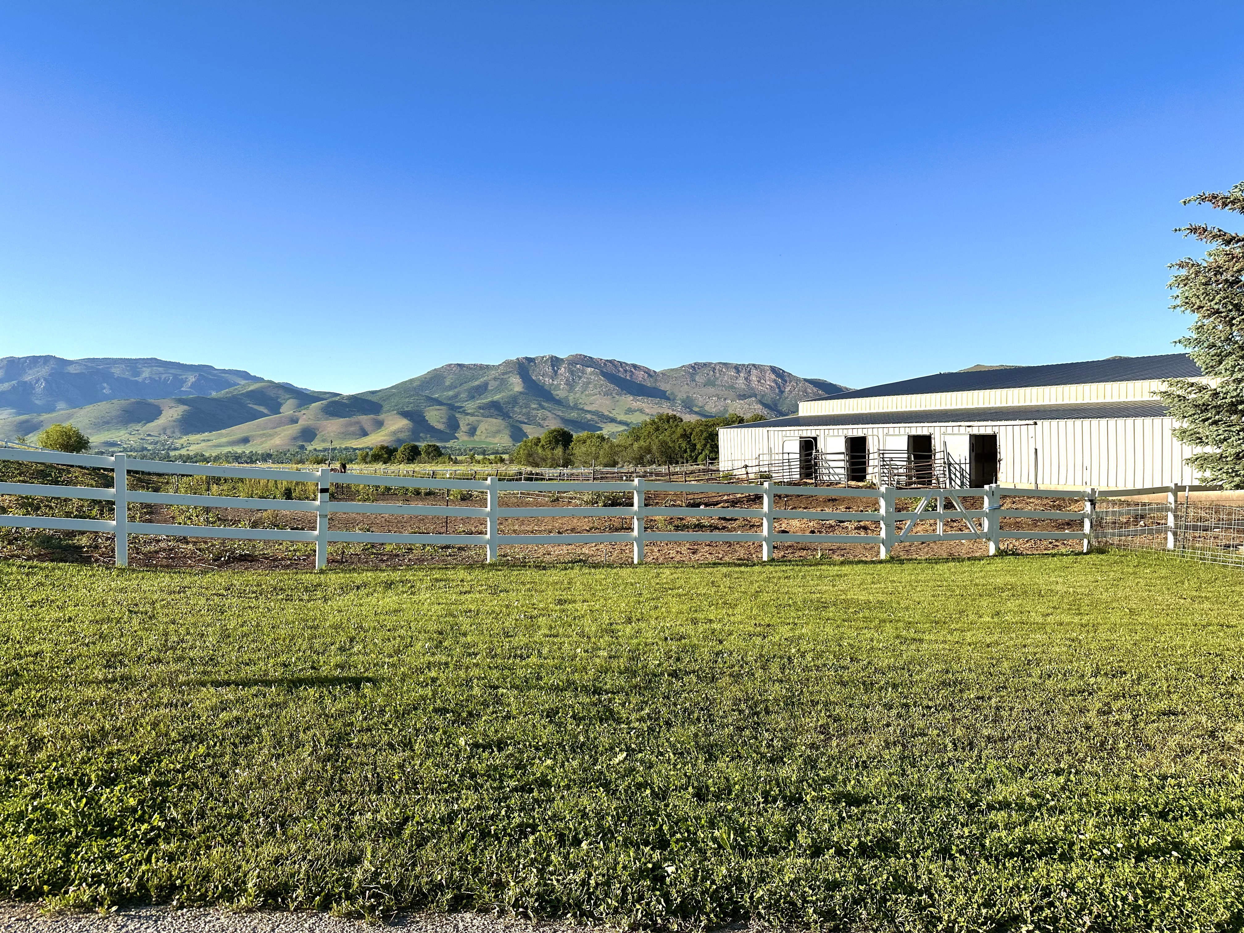 Expansive outdoor area featuring a white perimeter fence, a large metal-sided outbuilding with multiple stalls, and distant mountain scenery