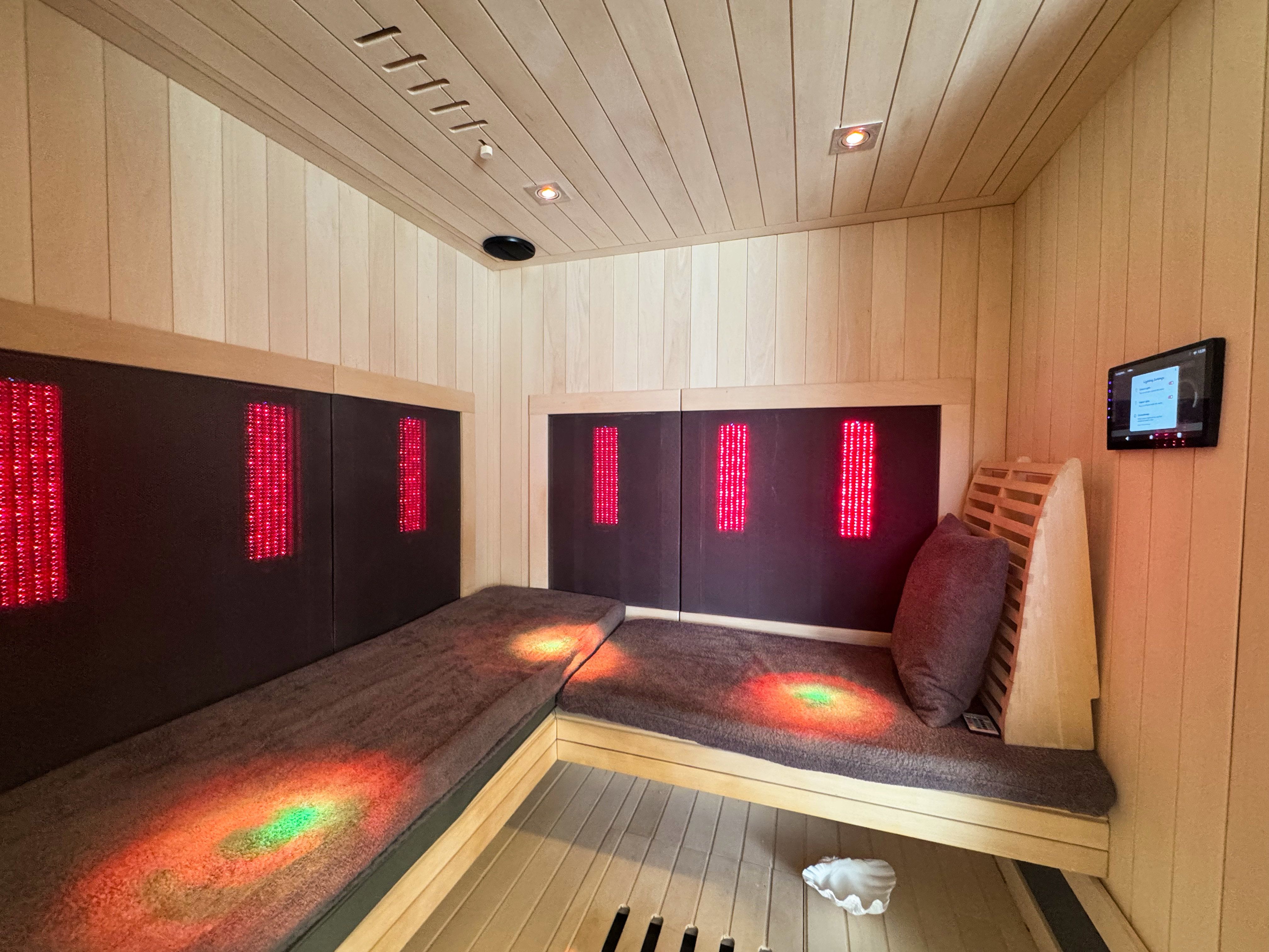 Custom-built sauna featuring light-toned wood-paneled walls and ceiling, built-in bench seating with plush cushions, integrated red light therapy panels, recessed ceiling lighting, and a wall-mounted control panel