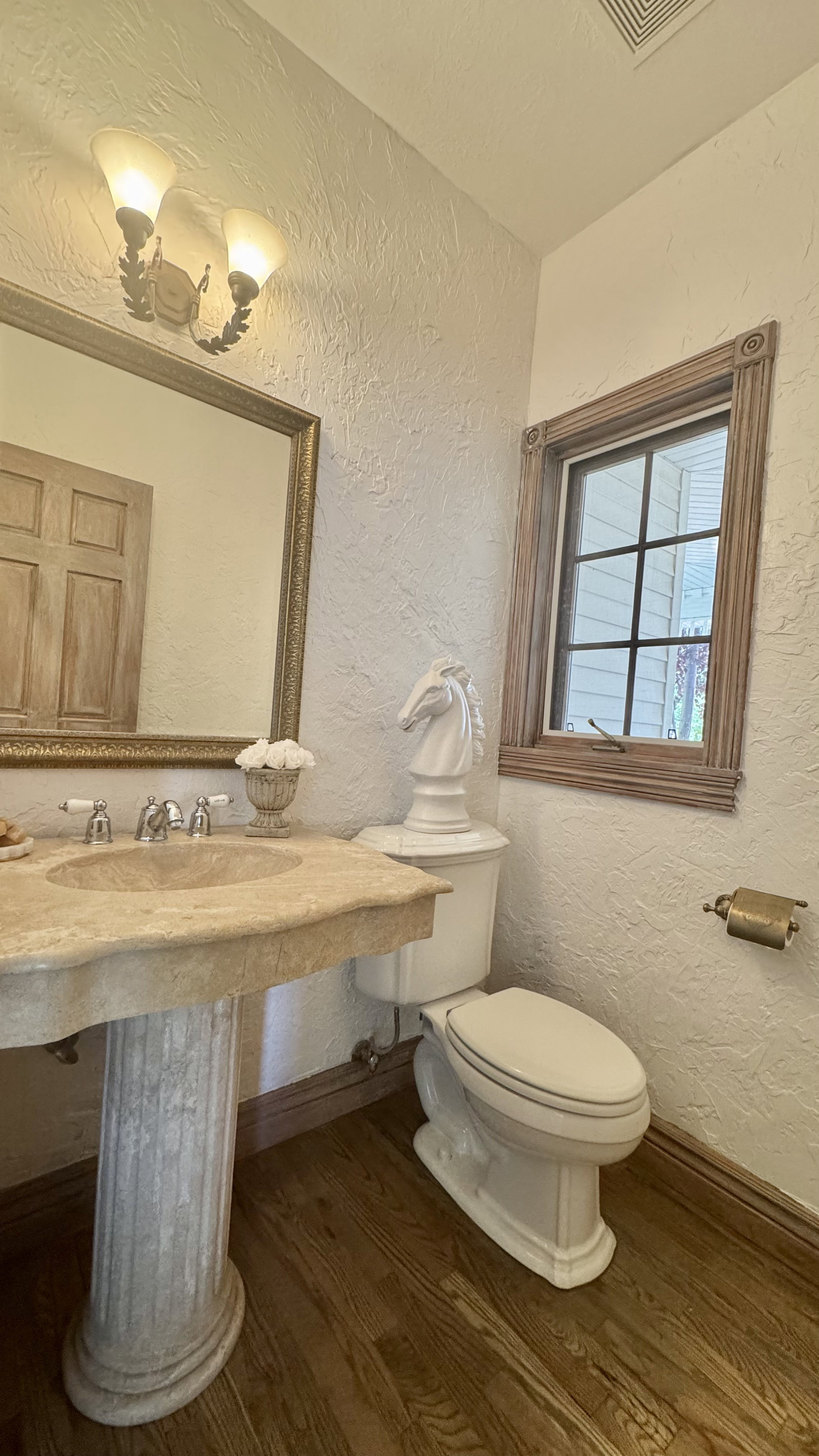 Pedestal sink with a stone finish, a wall-mounted light fixture, wood-finish flooring, a window with wood trim, and a white toilet