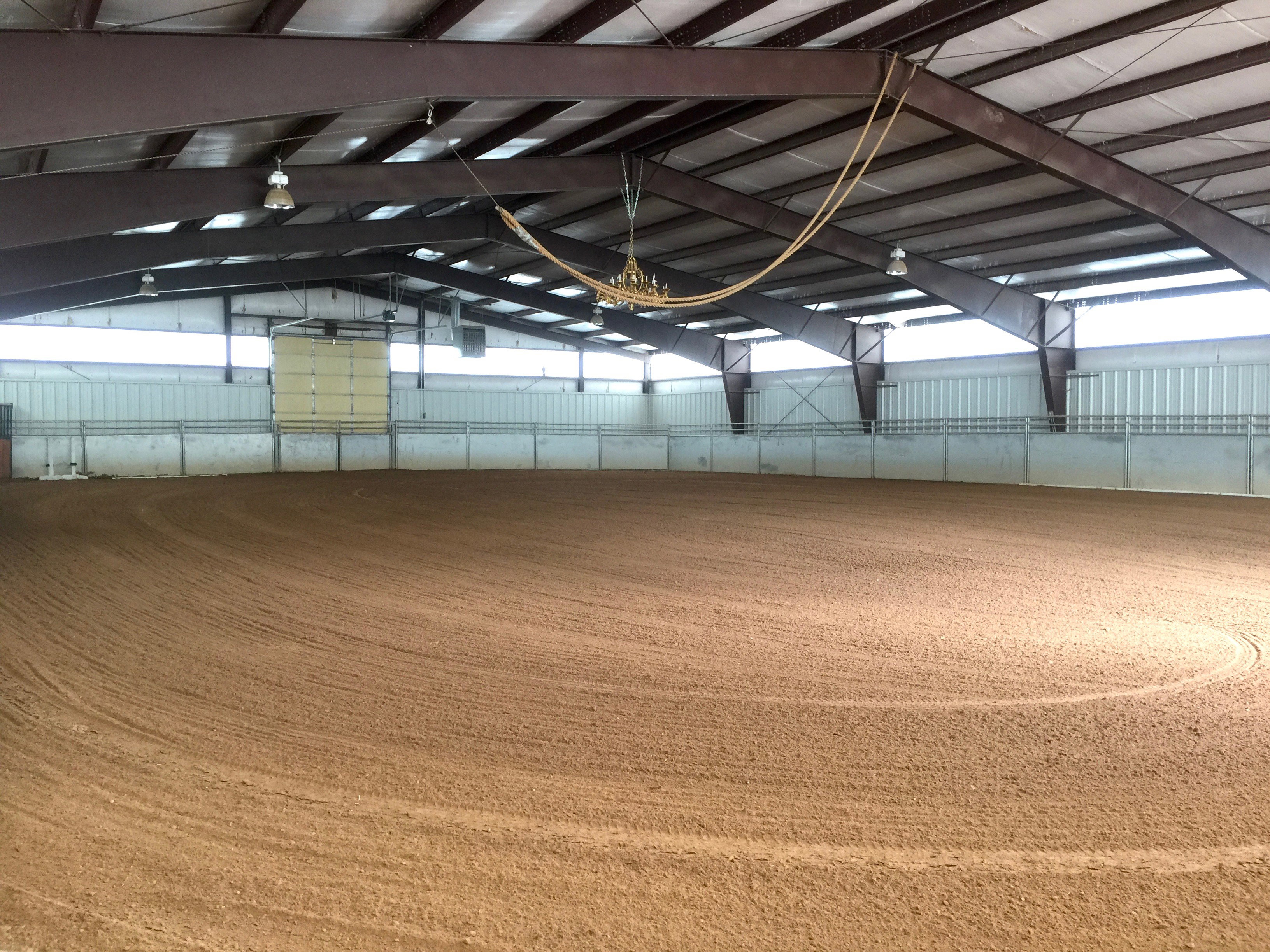 Spacious indoor arena featuring a sand footing, high ceilings with exposed steel beams, multiple overhead light fixtures, and a large roll-up door