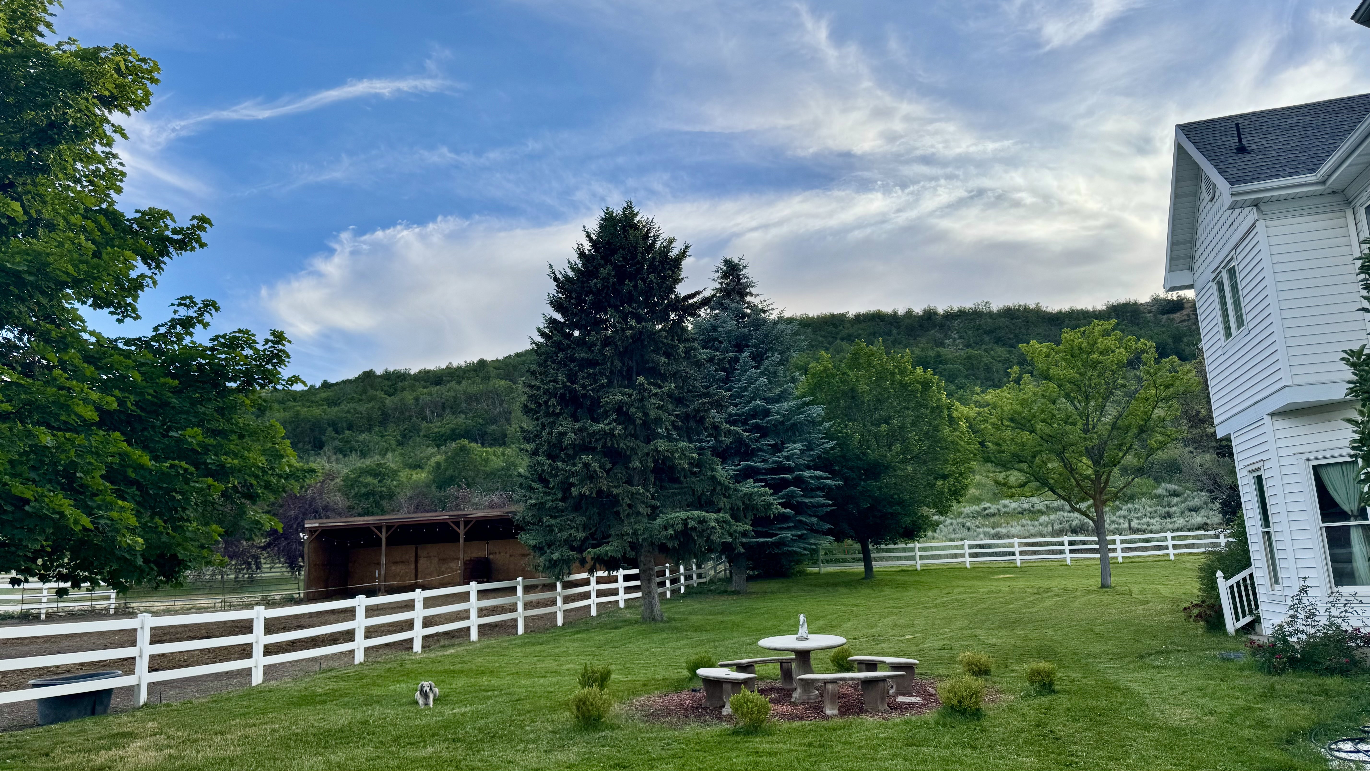 Expansive green lawn with a white fence, mature trees, and mountain scenery