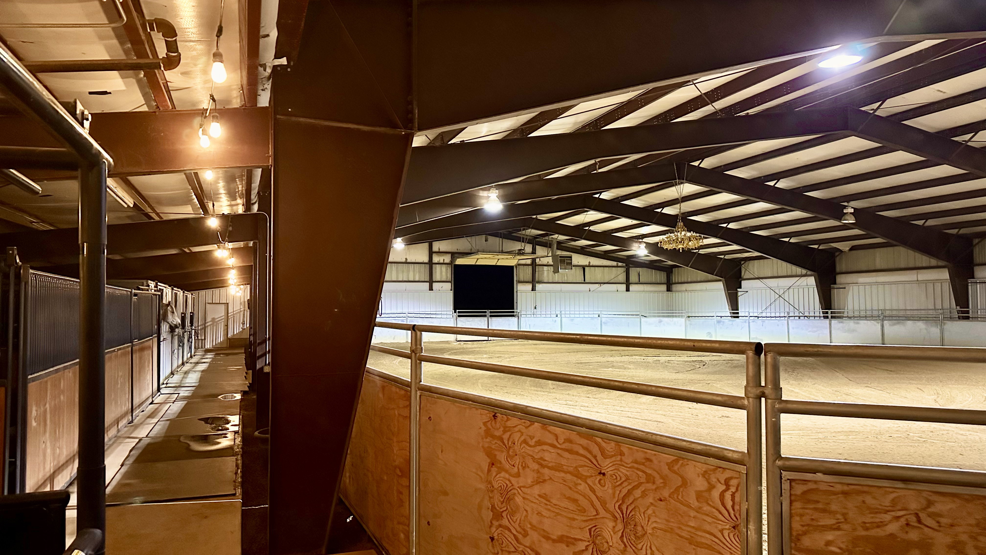 Indoor riding arena featuring overhead lighting, a sand footing surface, and a substantial steel-frame structure