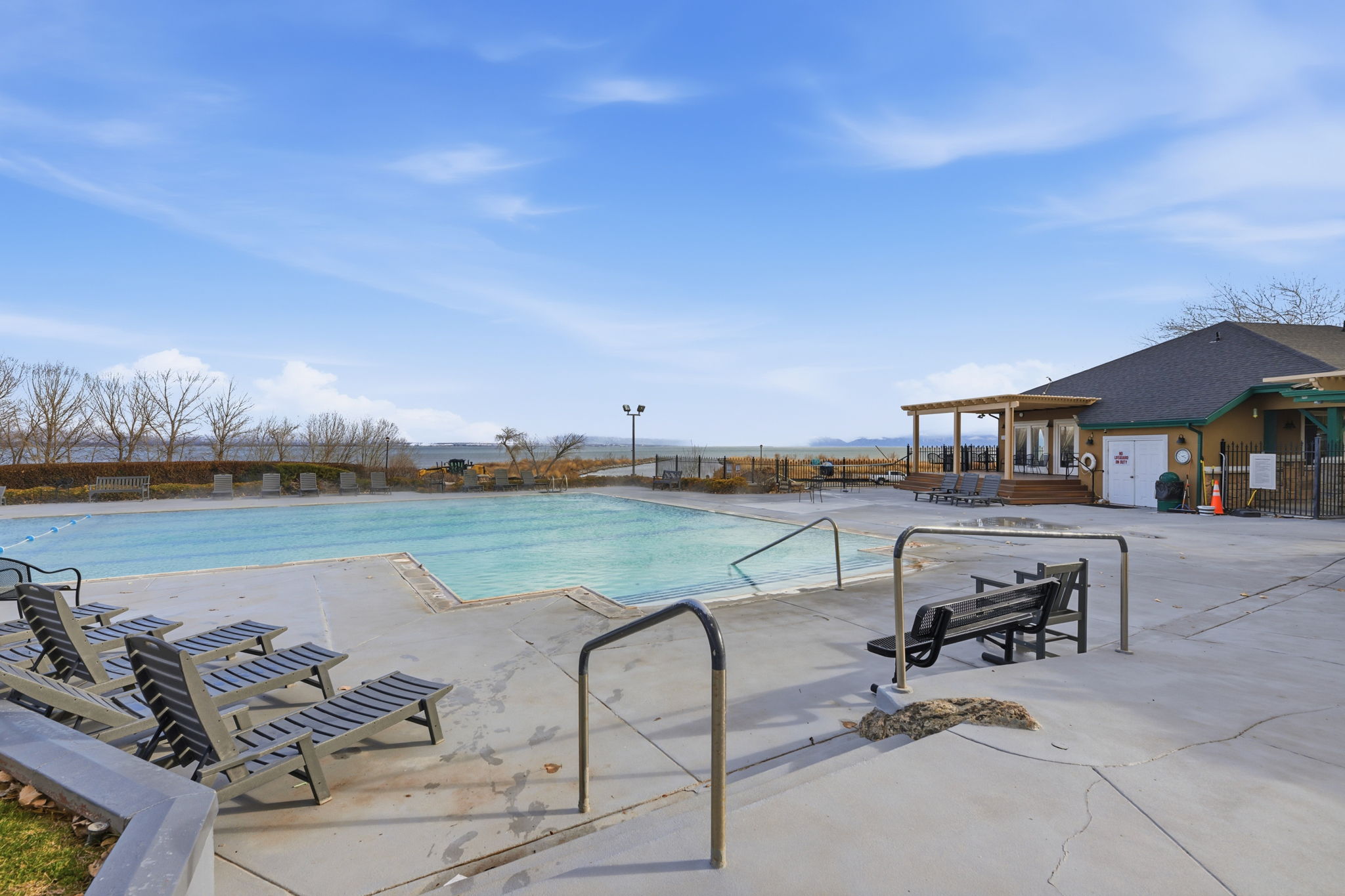 Outdoor swimming pool with integrated lane lines and concrete decking, featuring a surrounding fence, poolside lounge chairs, and a nearby building with a covered patio