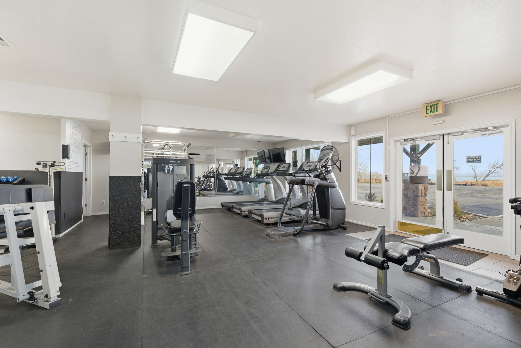 Expansive fitness center featuring large windows, reflective wall panels, recessed lighting, and durable rubber flooring