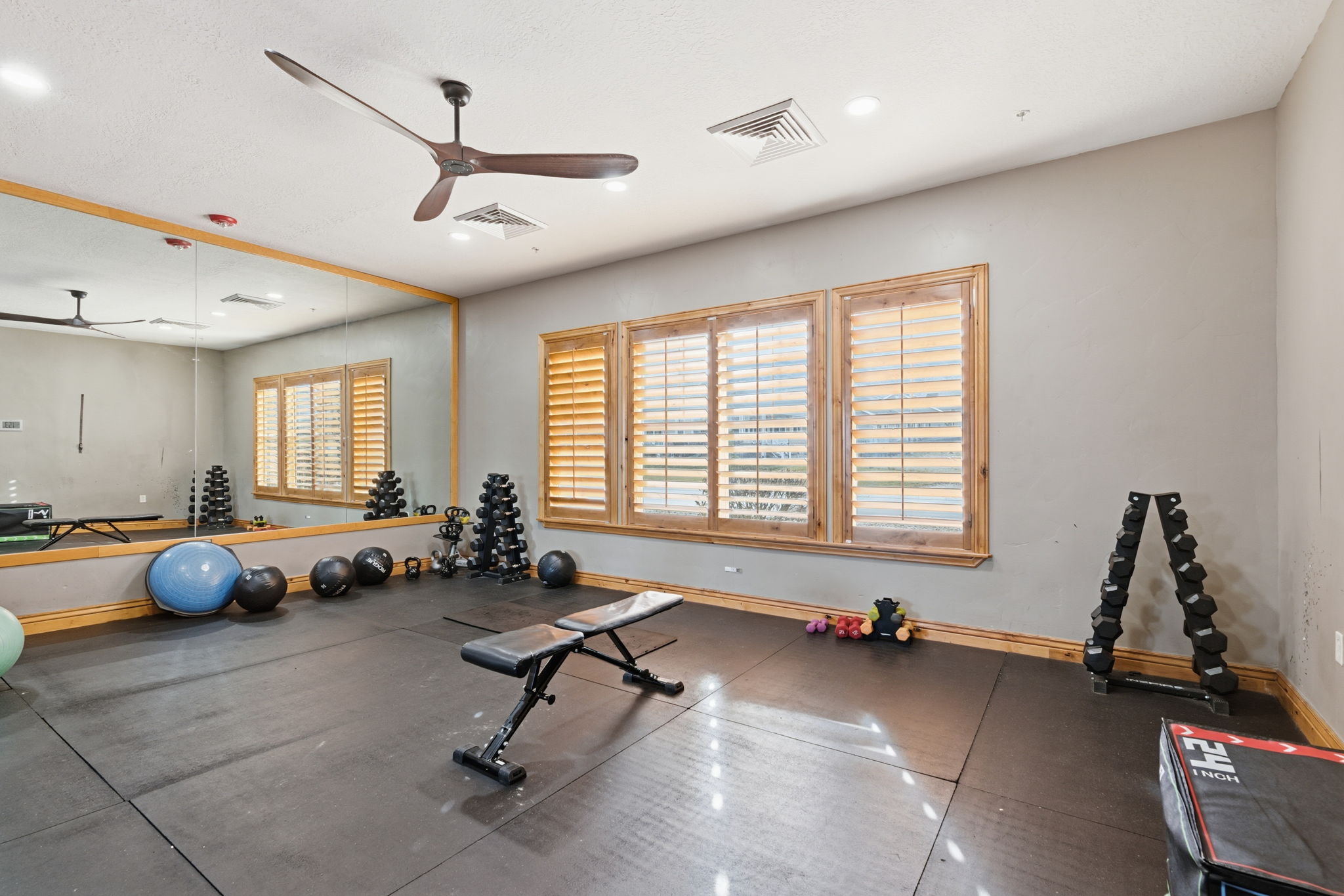 Dedicated fitness space featuring rubber flooring, a full-wall mirror, three windows with wood plantation shutters, a wood-finish ceiling fan, and recessed lighting