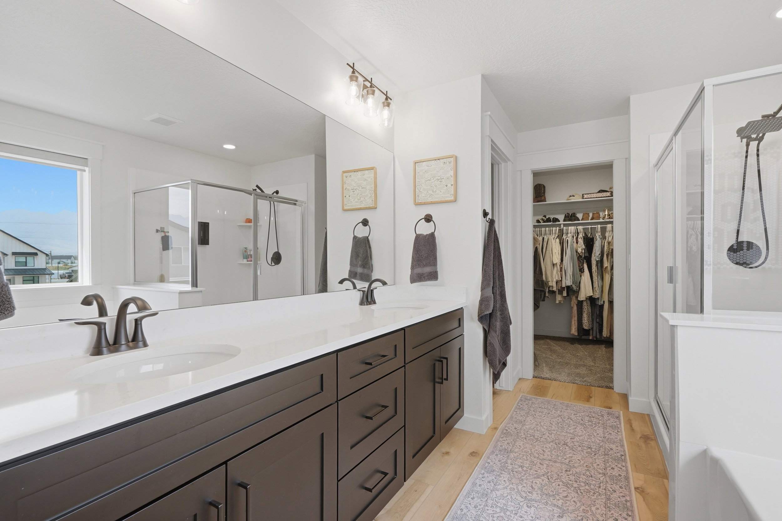 Bathroom featuring a dark wood double vanity with white countertops, bronze-finish fixtures, a large frameless mirror, and a glass-enclosed shower