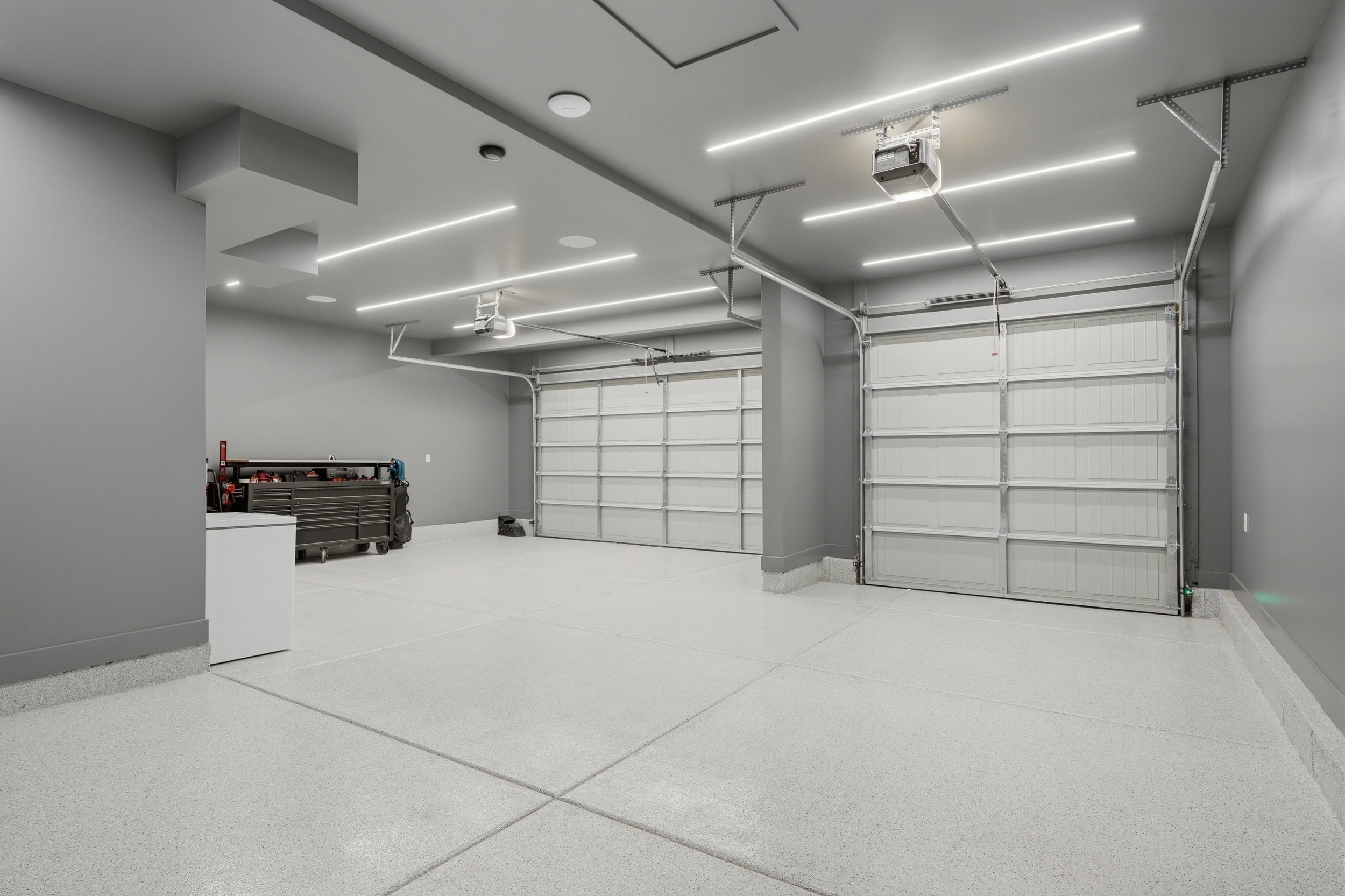Spacious garage featuring a light-colored epoxy floor, two garage doors with automatic openers, flush-mounted linear lighting, and gray painted walls
