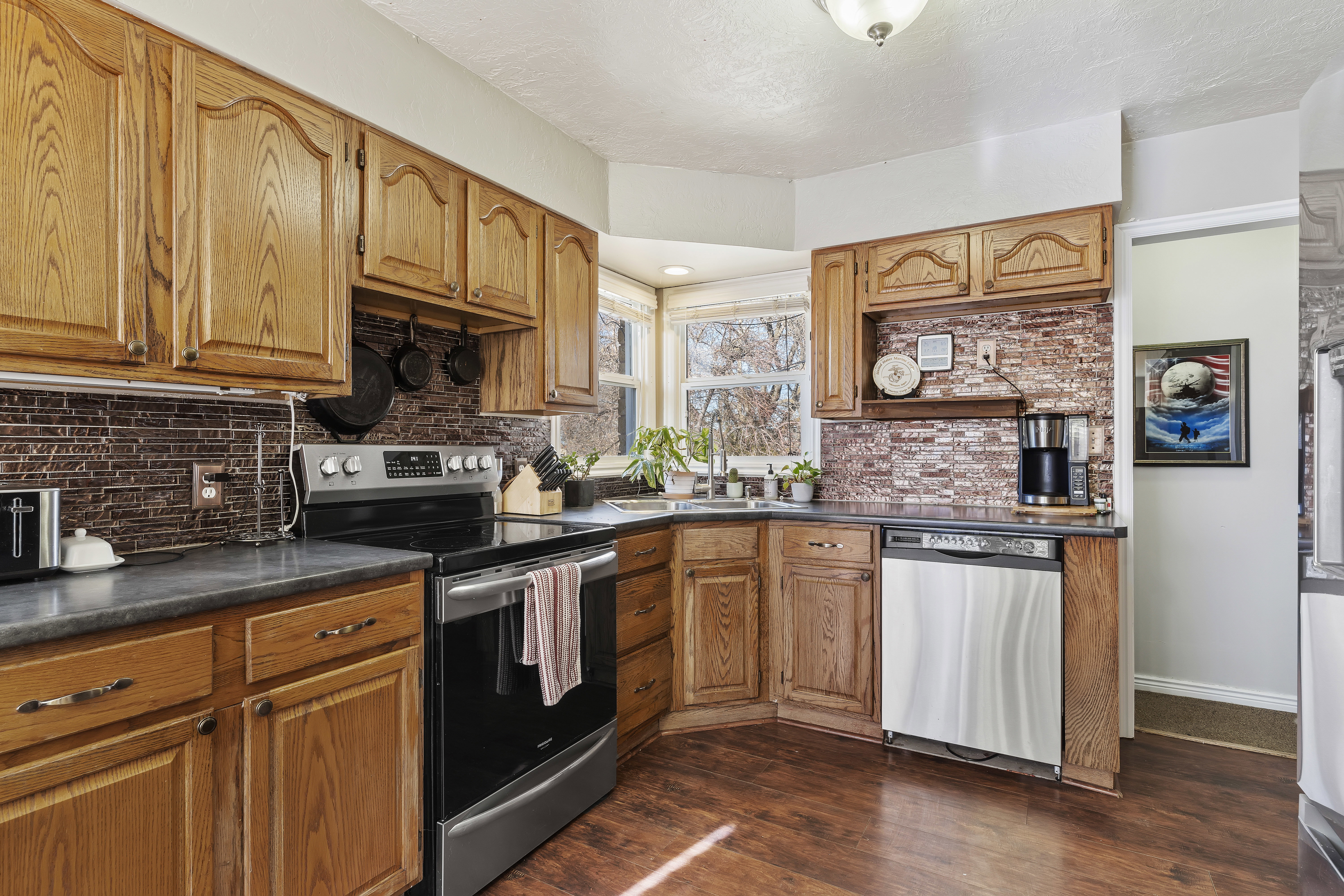 Kitchen featuring warm wood cabinetry, dark countertops, an integrated stainless steel dishwasher, a black and stainless steel range, and a textured brick-patterned backsplash