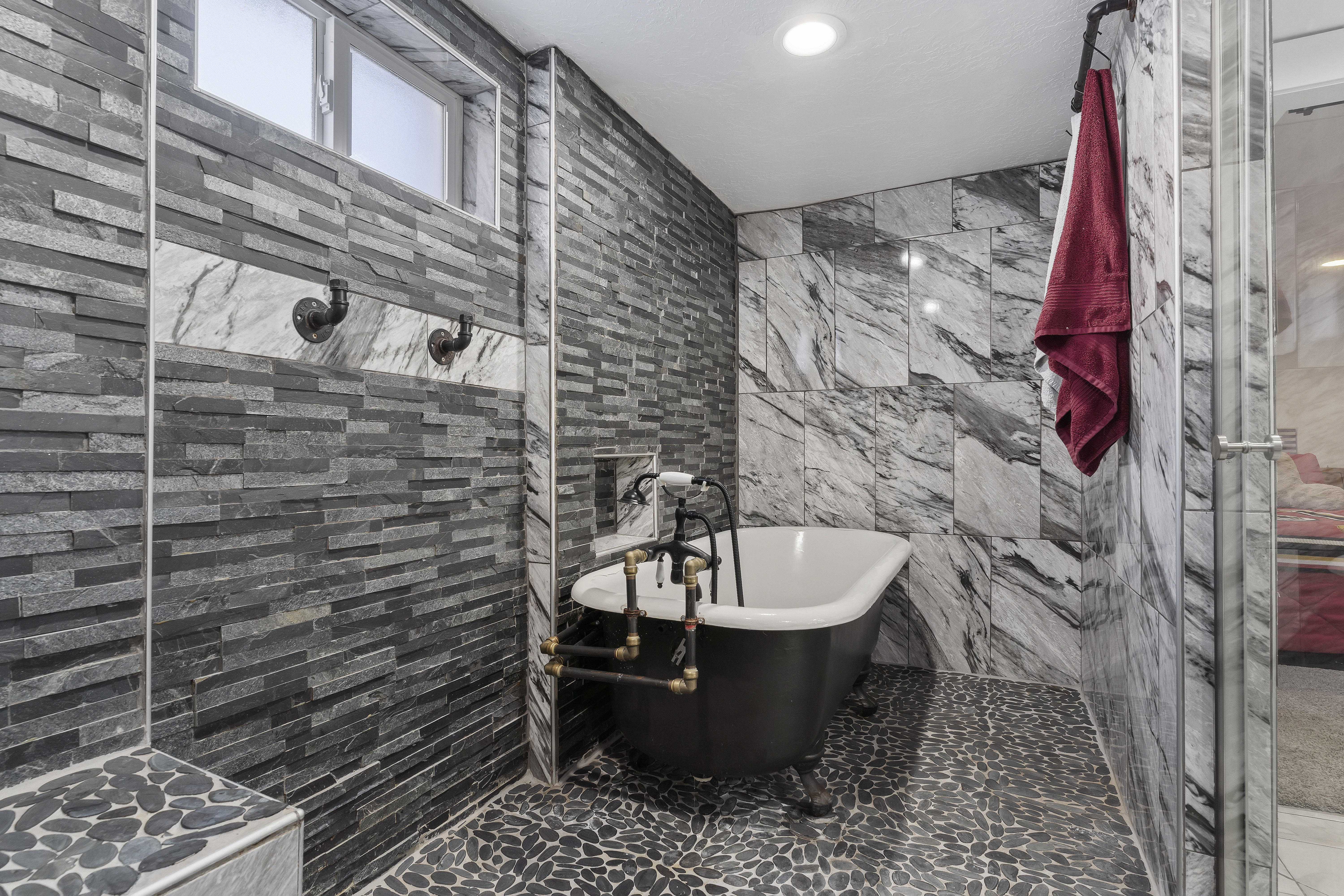Freestanding clawfoot tub with exposed pipe-style fixtures, set against a backdrop of varied stone and tile finishes