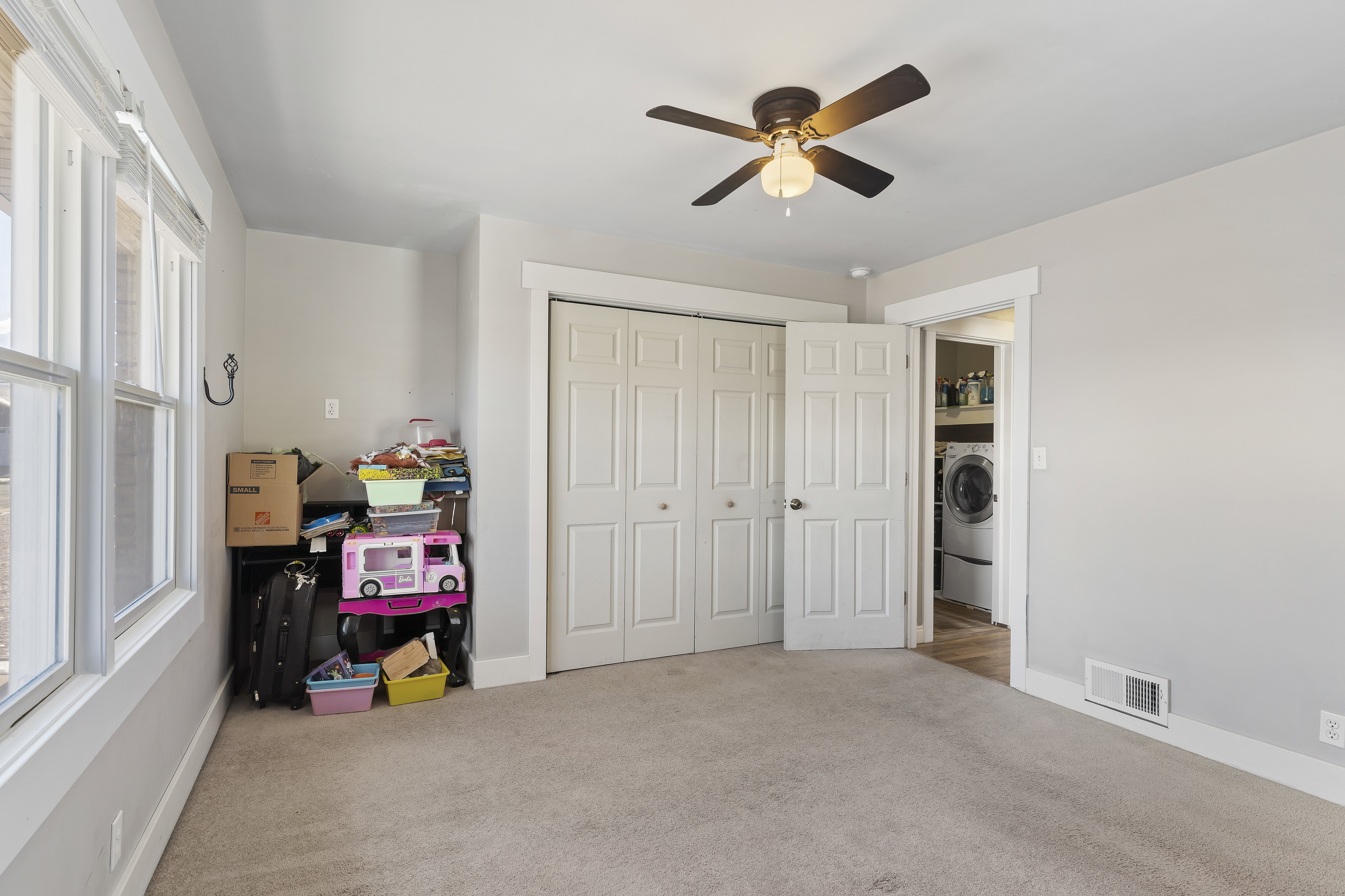 Carpeted room featuring a ceiling fan, dual windows, bi-fold closet doors, and access to an in-unit laundry area with a visible washer