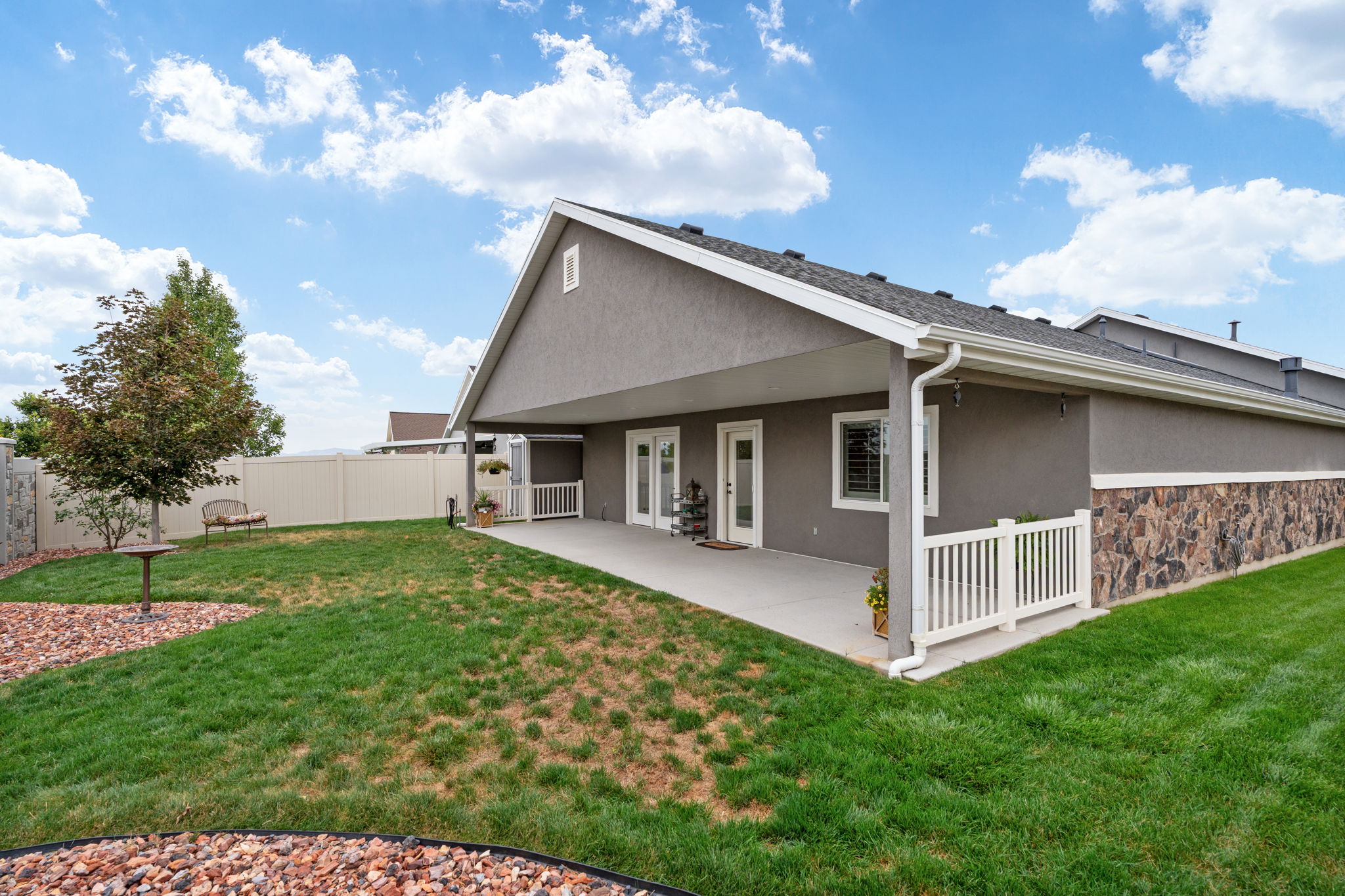 Expansive backyard featuring a covered patio, stucco exterior, stone accent detailing, a white vinyl fence, and a mature tree