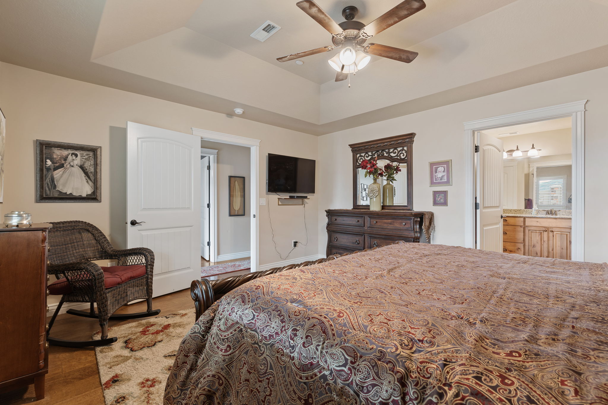 Tray ceiling bedroom with wood-finish flooring, a ceiling fan, and direct access to an en-suite bathroom featuring a double vanity with stone countertops