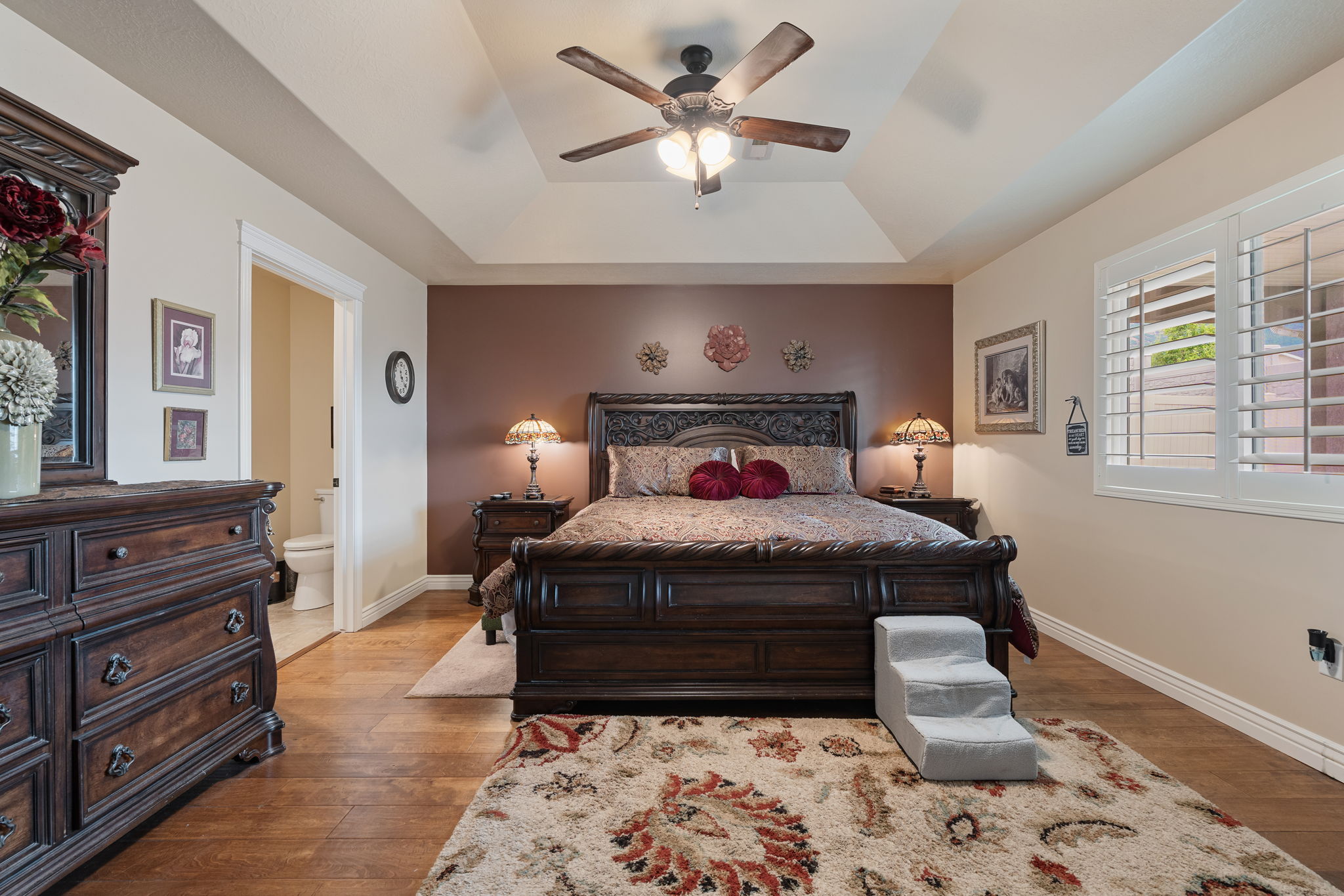 Spacious bedroom featuring wood-finish flooring, a tray ceiling with a ceiling fan, and white plantation shutters