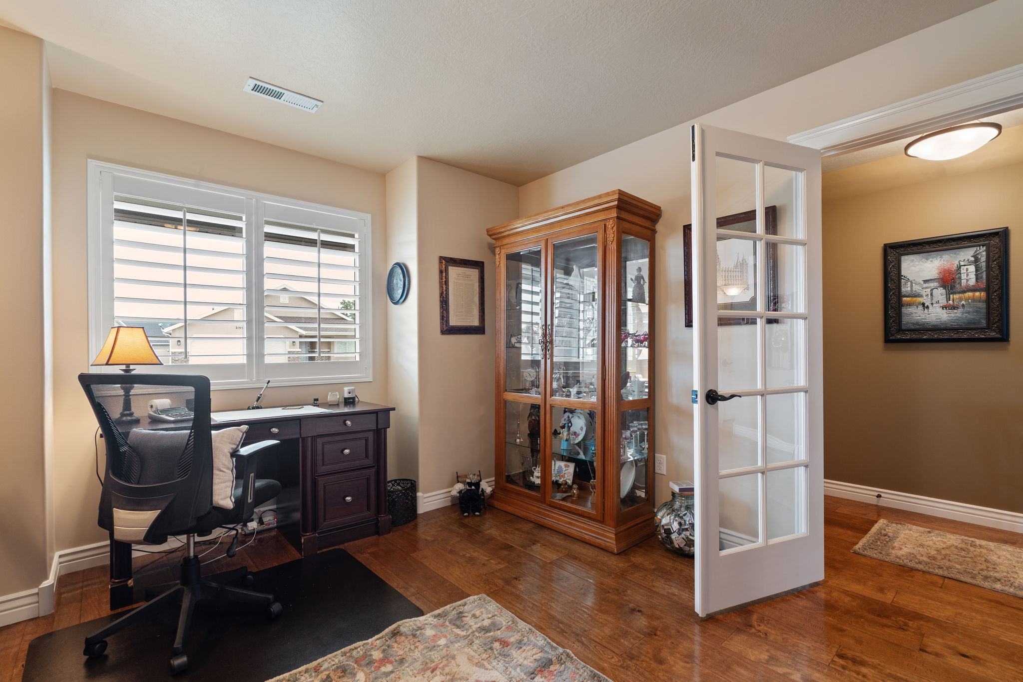 Hardwood flooring throughout this room, featuring a window with white plantation shutters, a glass-paneled interior door, crown molding, and a built-in ceiling light fixture