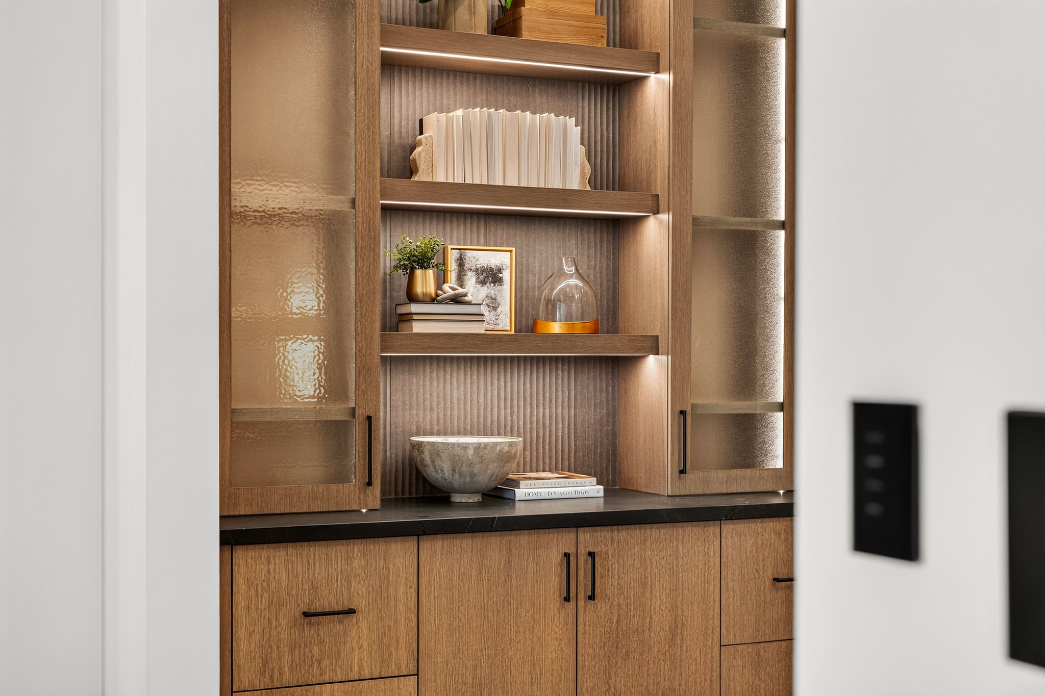 Integrated wood-finish cabinetry featuring open shelving with accent lighting, textured backing, and frosted glass cabinet doors