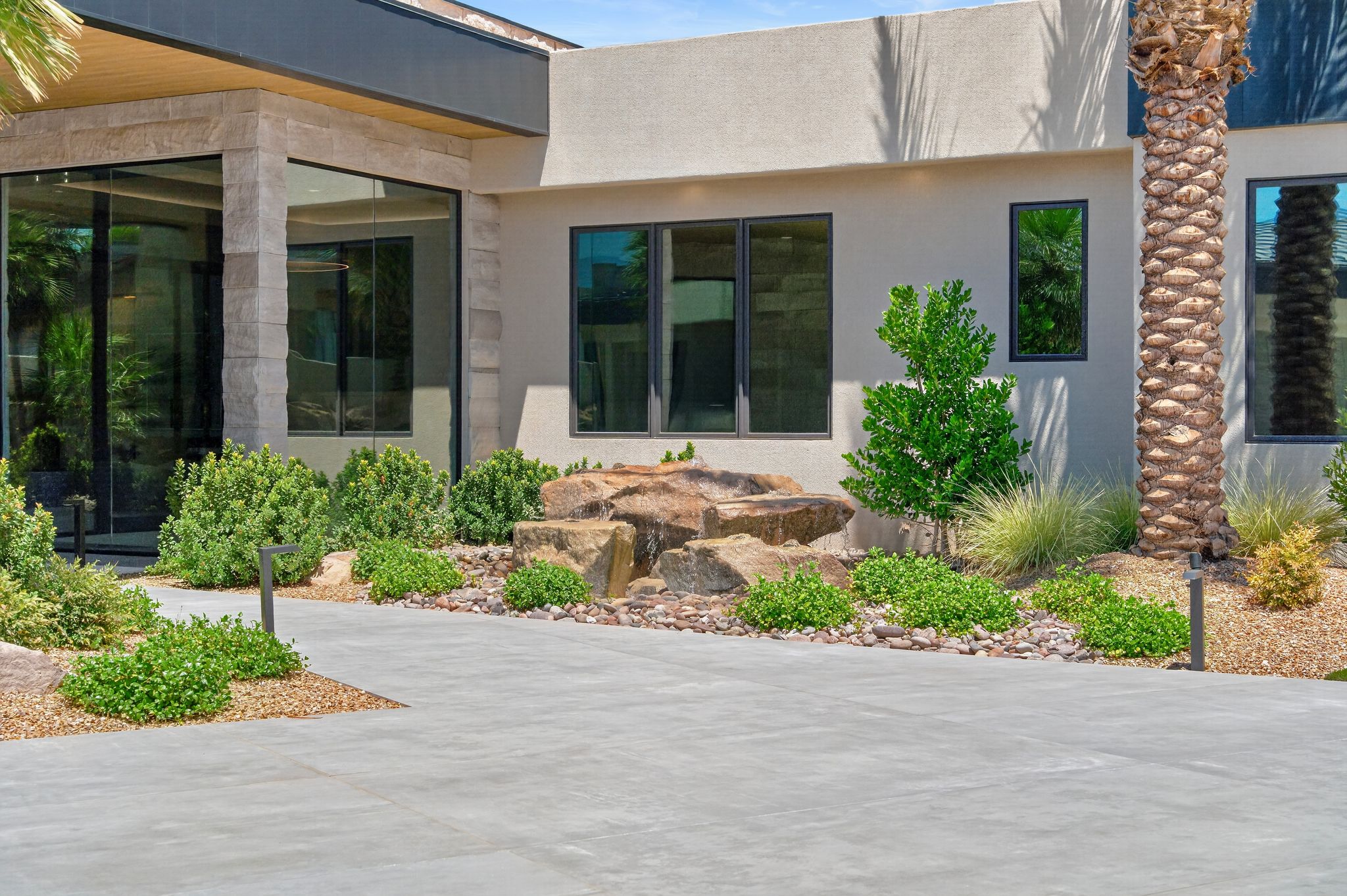 Modern exterior featuring stucco siding, stone accents, expansive black-framed windows, and mature desert landscaping with accent lighting
