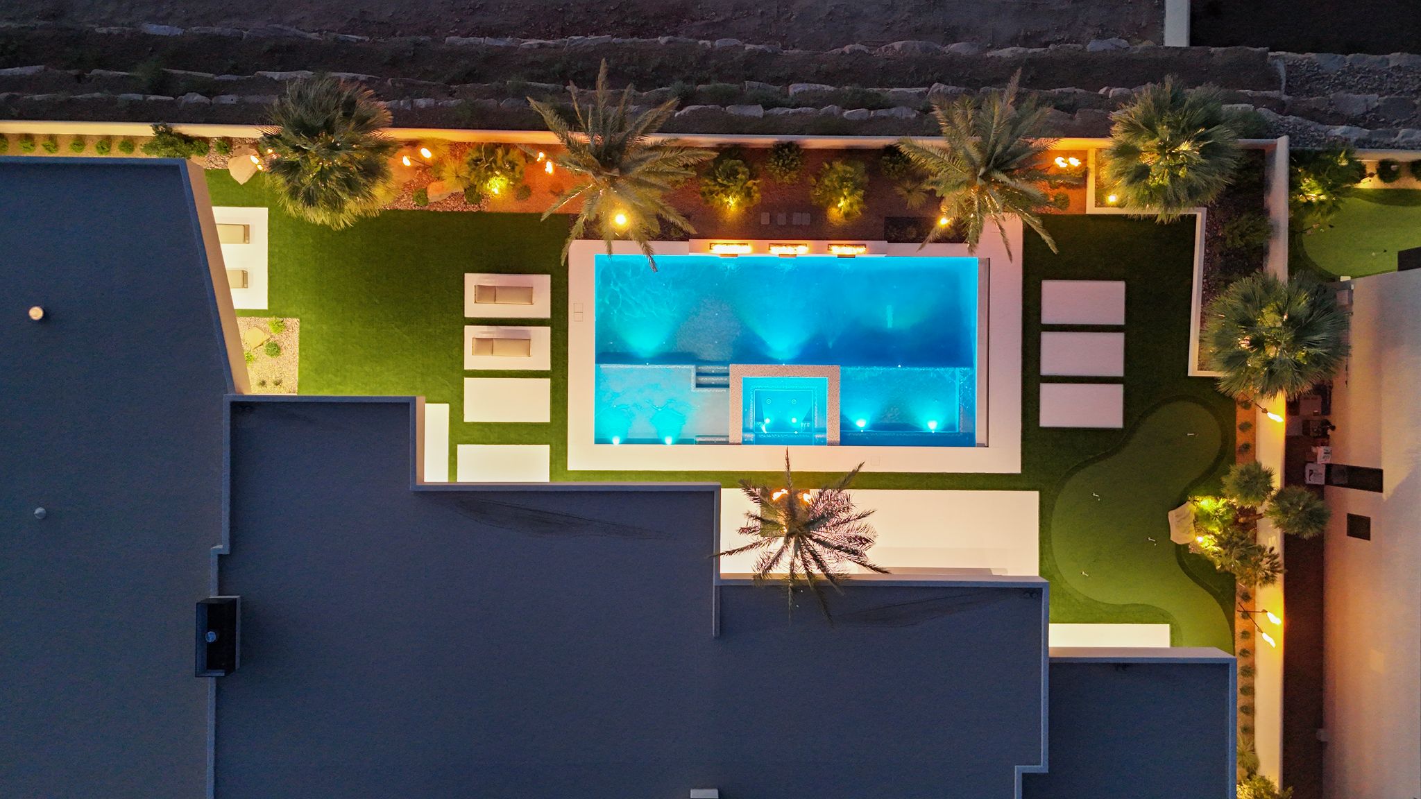 Backyard oasis featuring a rectangular swimming pool with integrated spa and lighting, extensive artificial turf, a putting green, and landscaped borders with mature palms