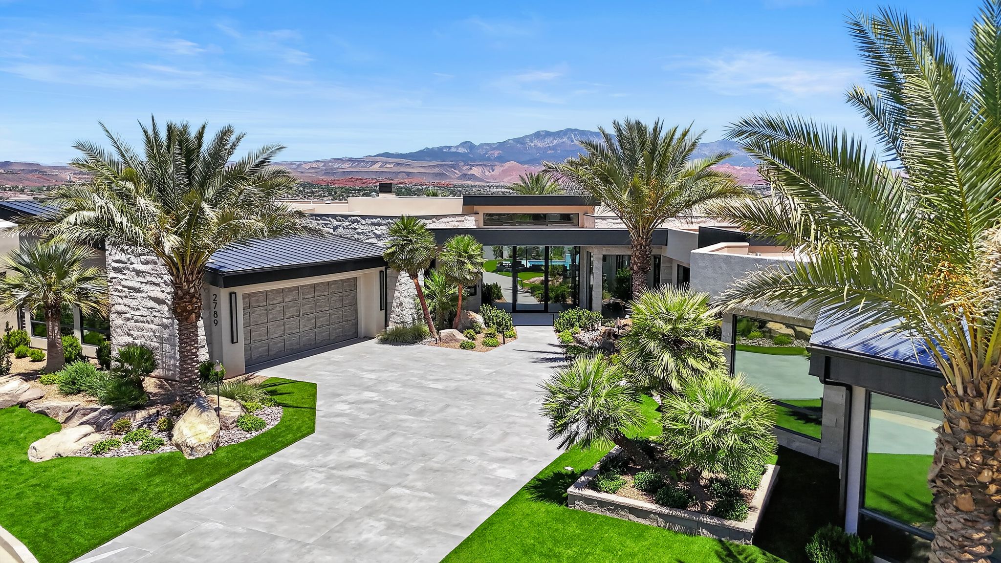 Modern architectural residence featuring extensive stone veneer, expansive glass walls, a textured garage door, a concrete driveway, and mature palm trees