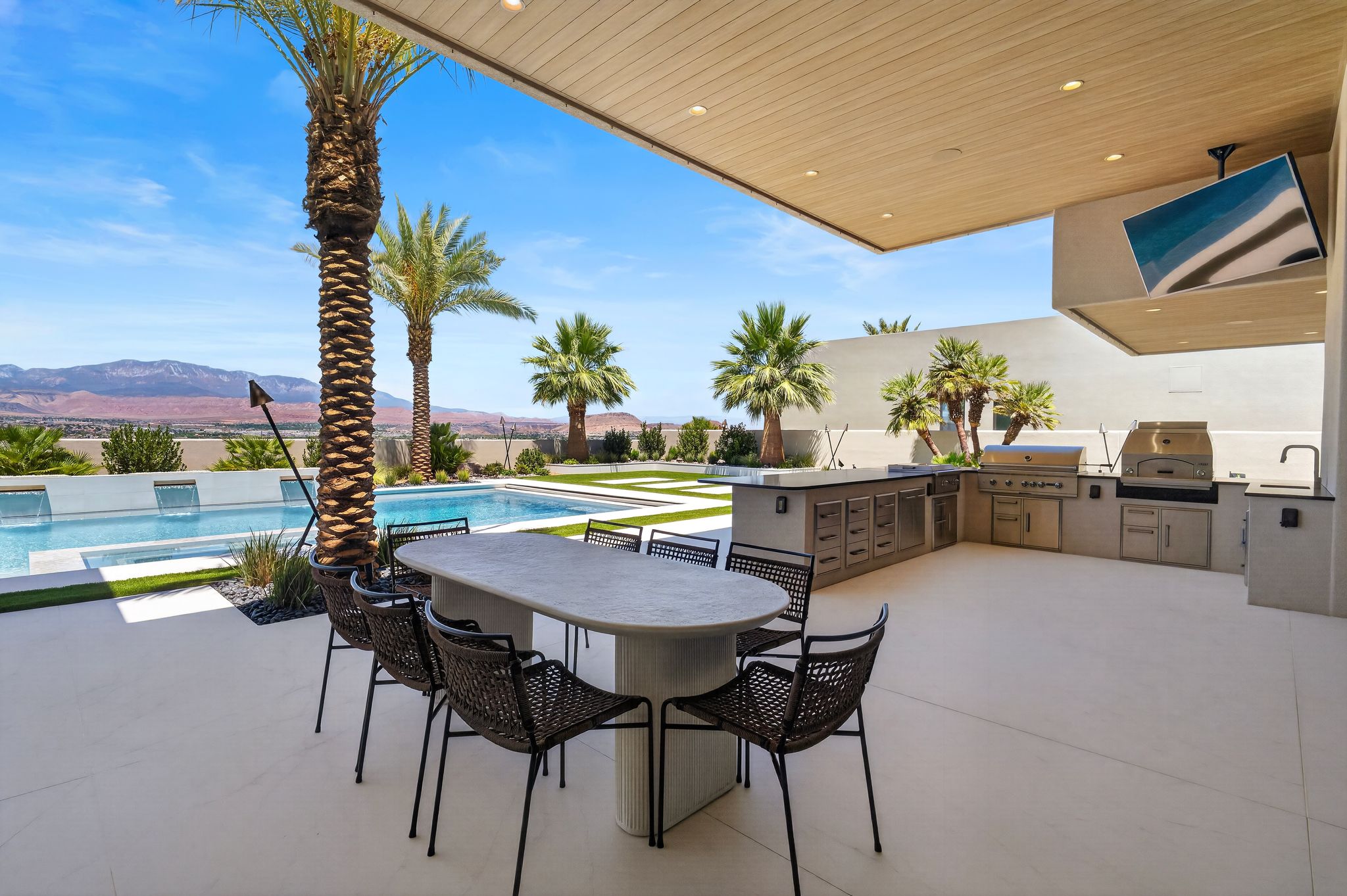 Expansive outdoor living area featuring a covered patio, built-in outdoor kitchen with stainless steel appliances, swimming pool with water features, and desert mountain scenery