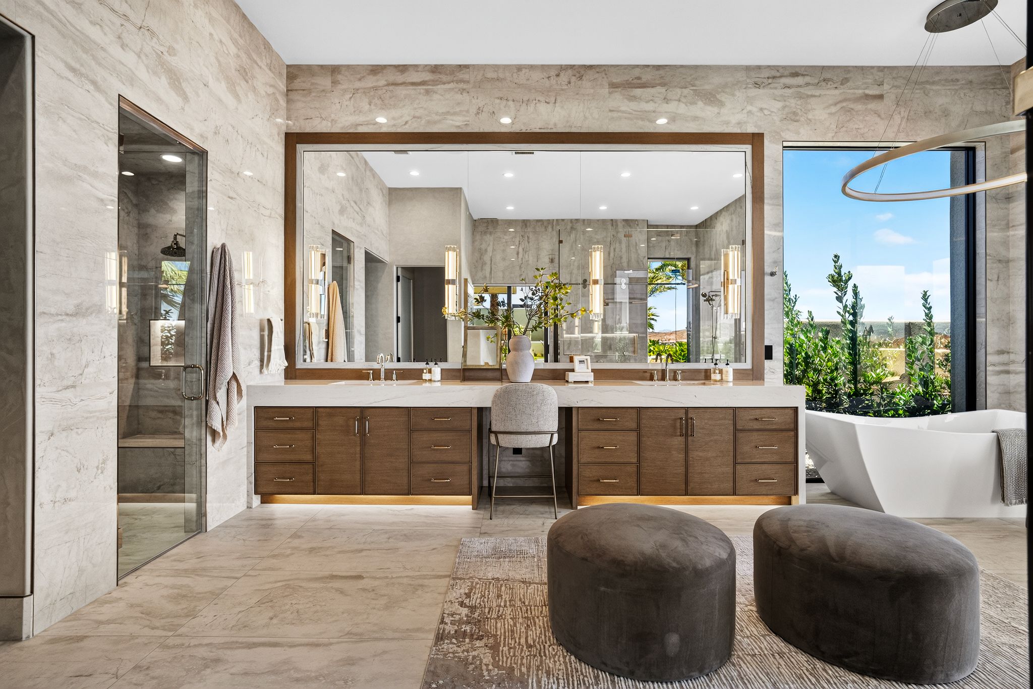 Spacious bathroom featuring a large frameless mirror, dual wood-finish vanity with stone countertop, freestanding soaking tub, and a glass-enclosed shower with a rainfall showerhead