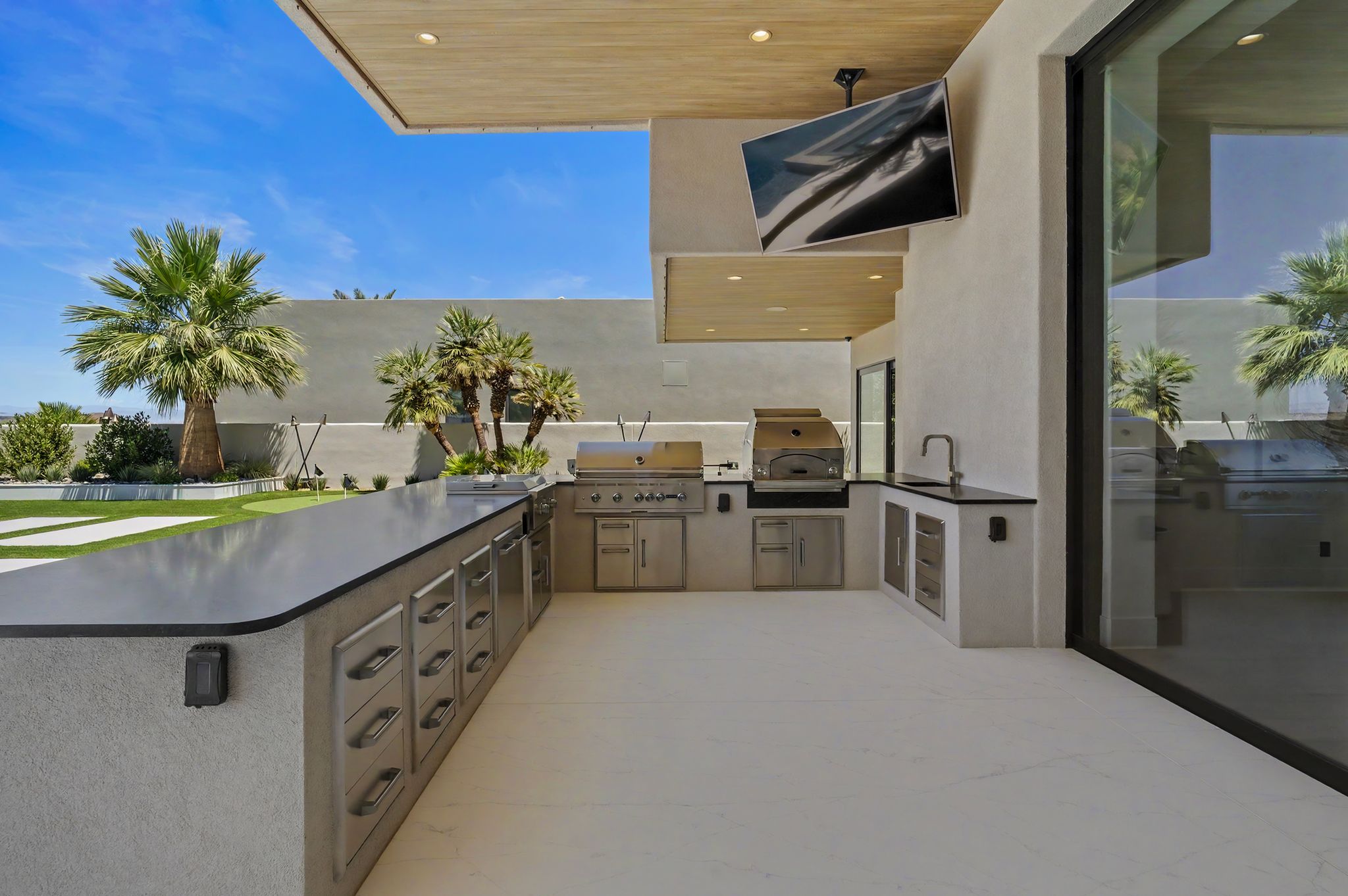 Outdoor kitchen featuring extensive countertop space, built-in stainless steel appliances, a sink with a gooseneck faucet, a ceiling-mounted television, and a wood-finish ceiling