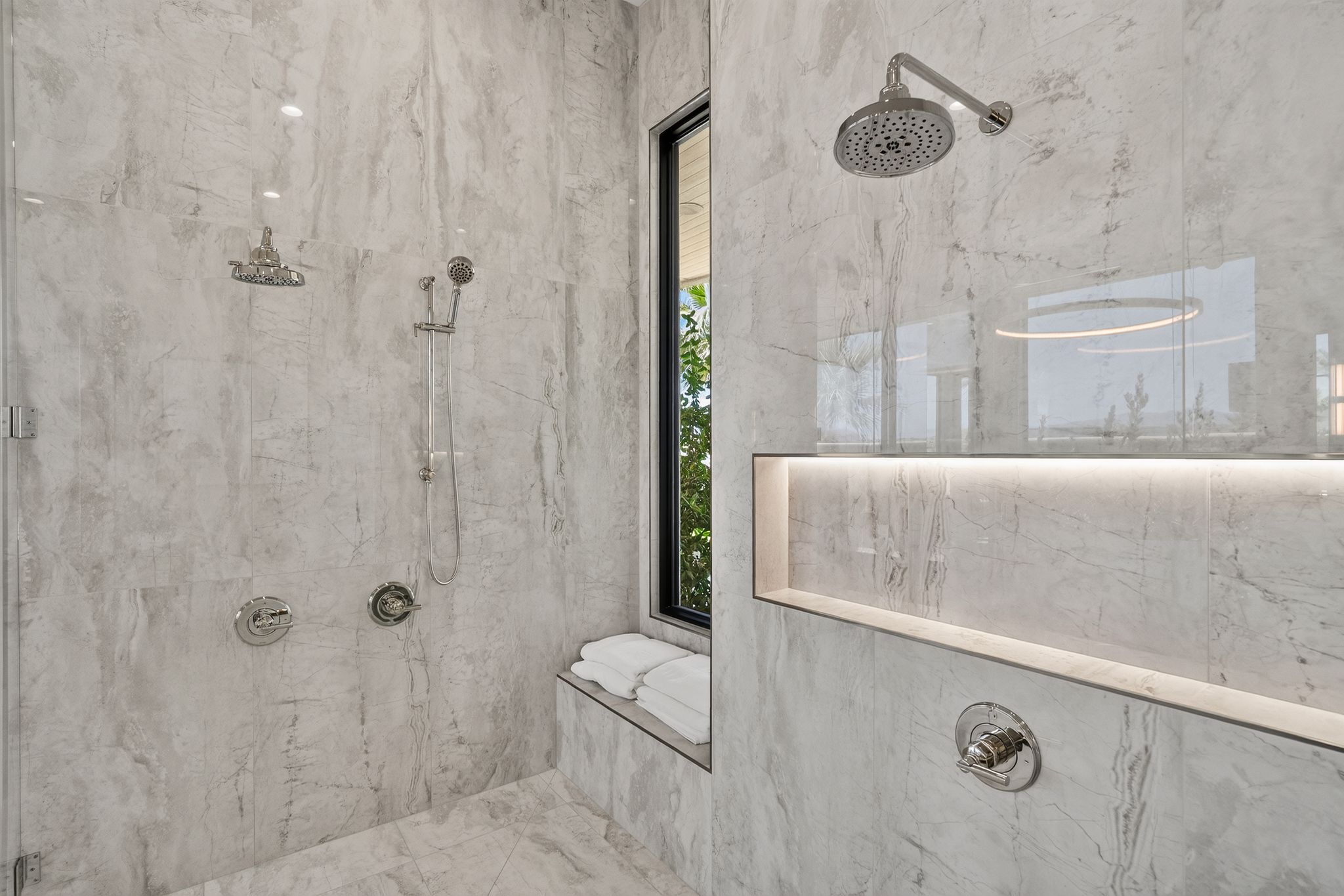 Spacious shower featuring light-toned stone-look tile, dual showerheads including a handheld sprayer, a built-in bench, a recessed niche with integrated lighting, and a black-framed window