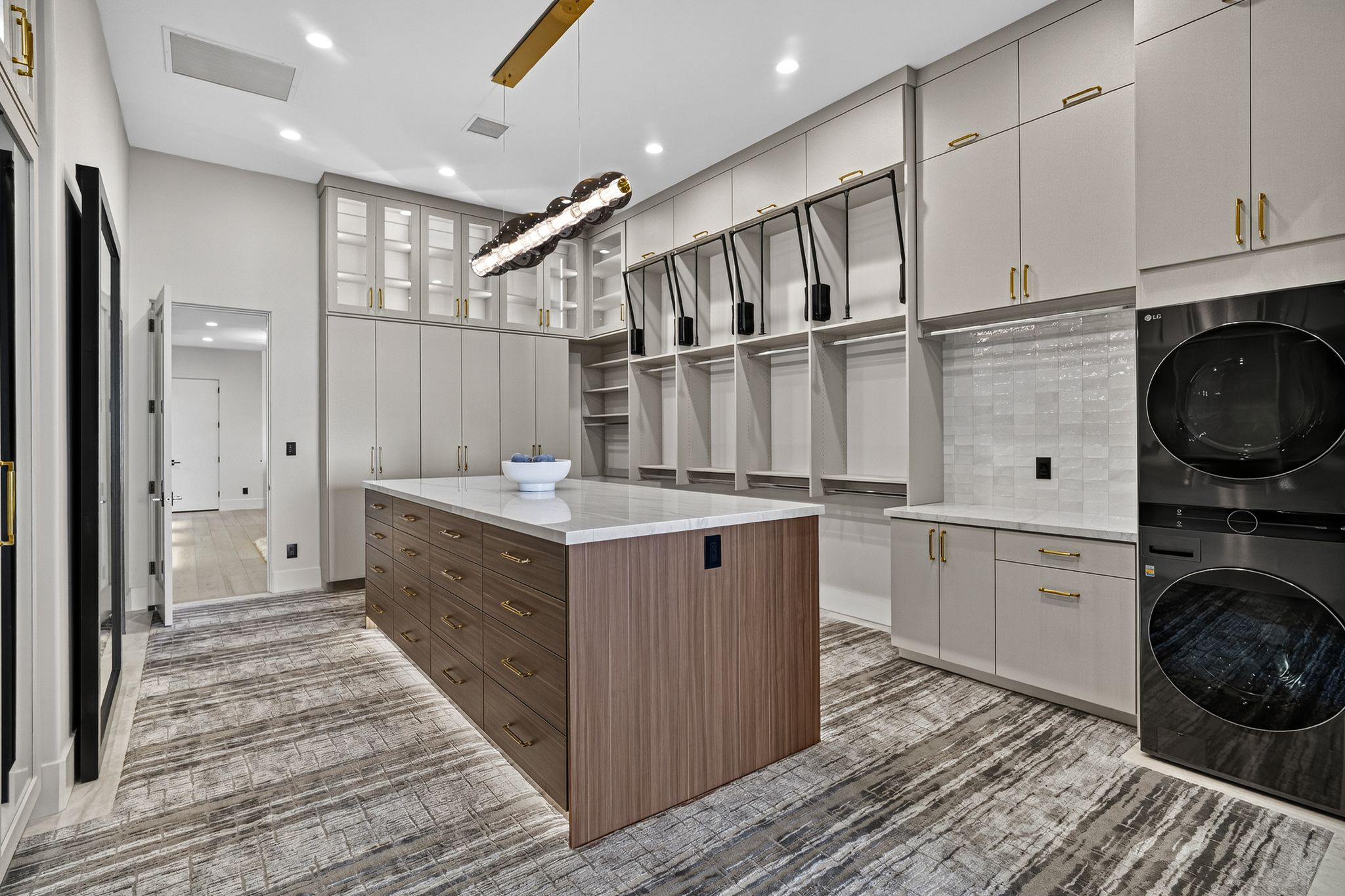 Spacious dressing room featuring a central island with a light-toned countertop, extensive built-in cabinetry with gold-toned hardware, and integrated laundry appliances