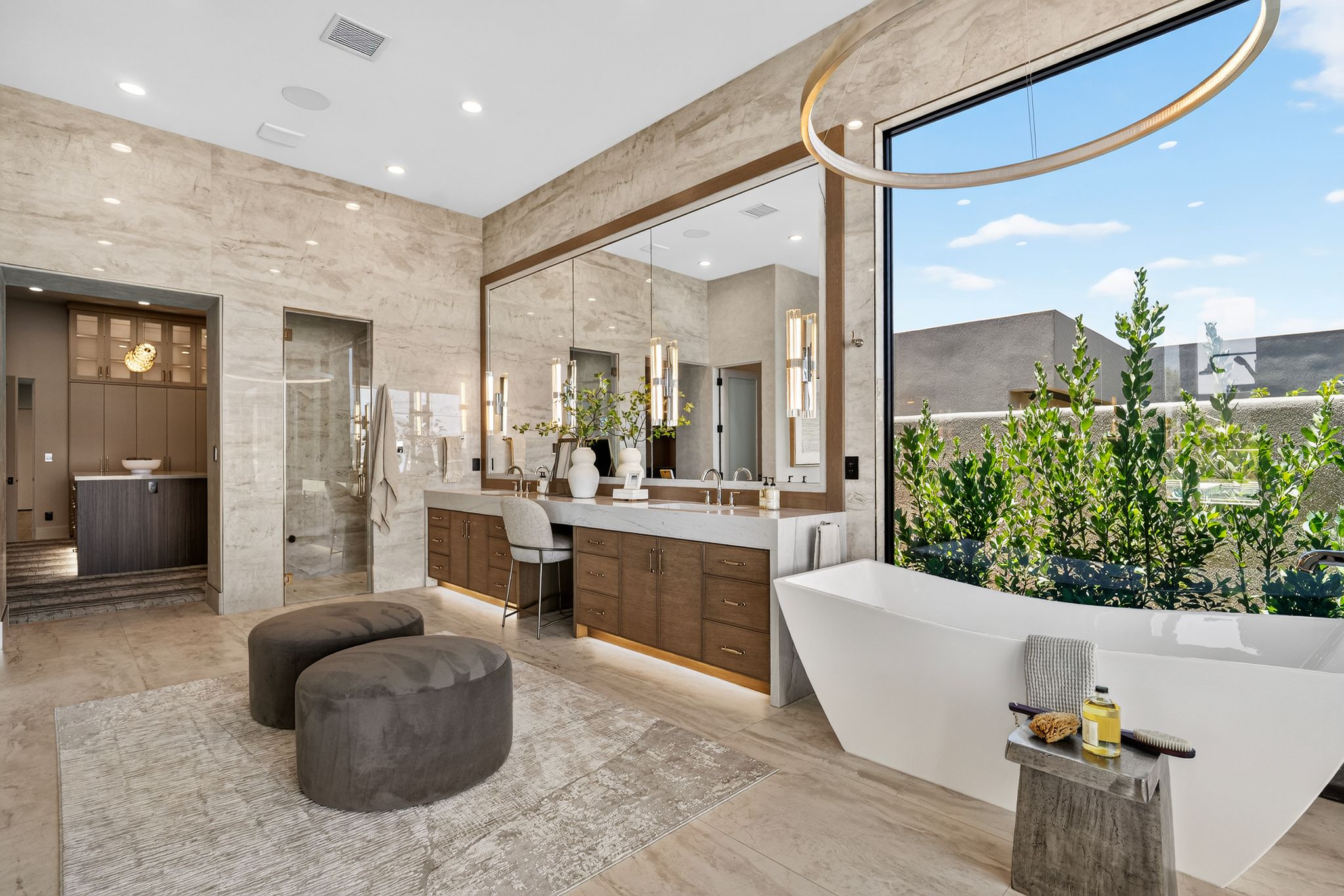 Contemporary bathroom featuring a freestanding soaking tub, expansive window, stone tile flooring and walls, and a wood-finish double vanity with integrated lighting