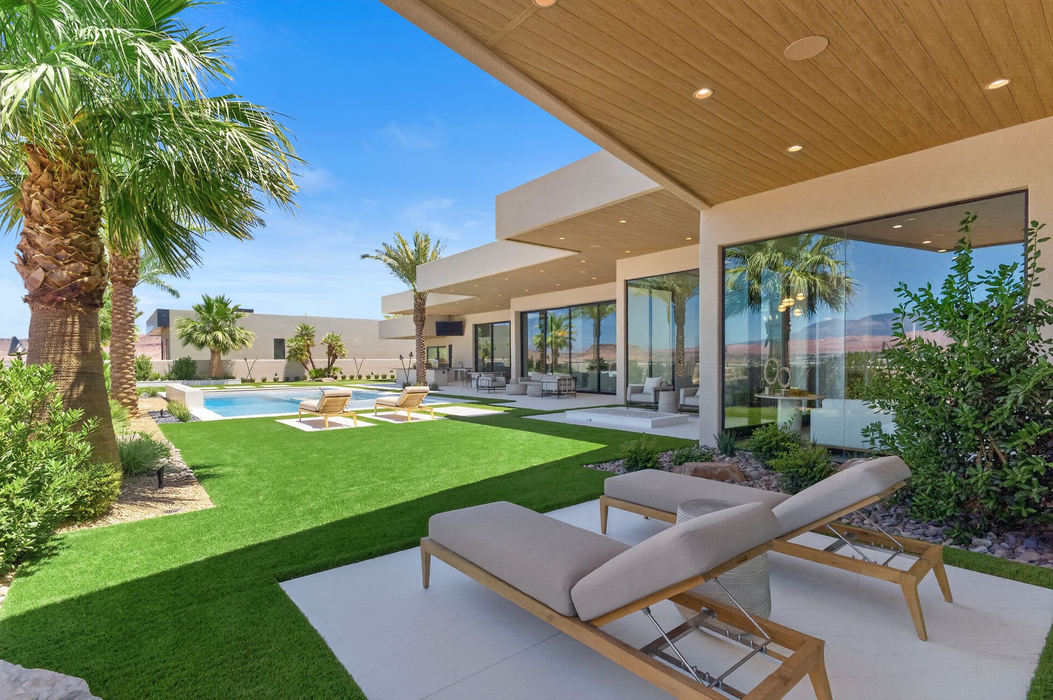 Modern exterior with expansive glass walls, a covered patio featuring a wood-finish ceiling, an artificial turf lawn, a built-in swimming pool, and mature palm trees