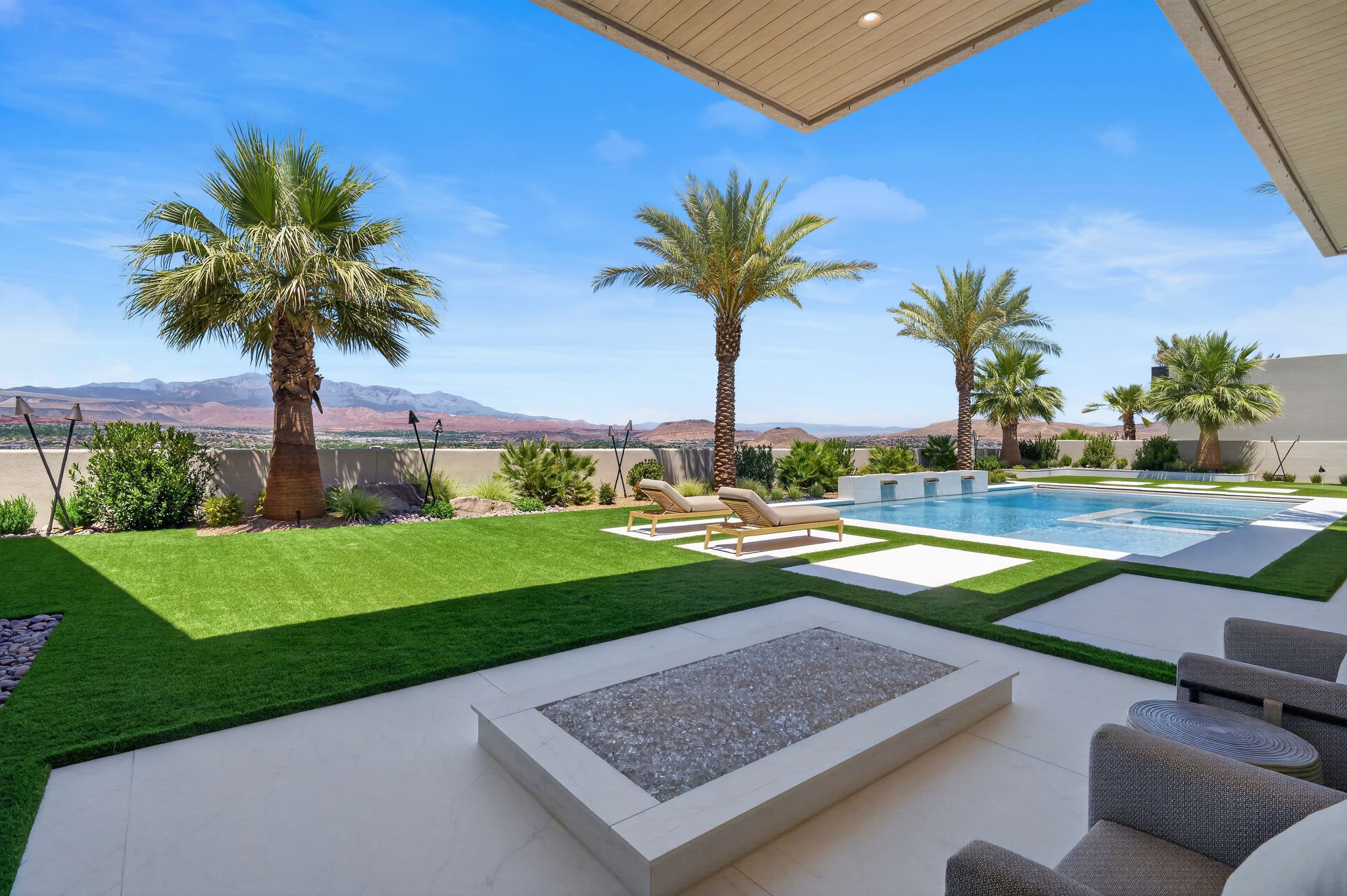 Expansive outdoor living area featuring a geometric swimming pool, integrated fire pit, artificial turf, mature palm trees, and a distant mountain landscape