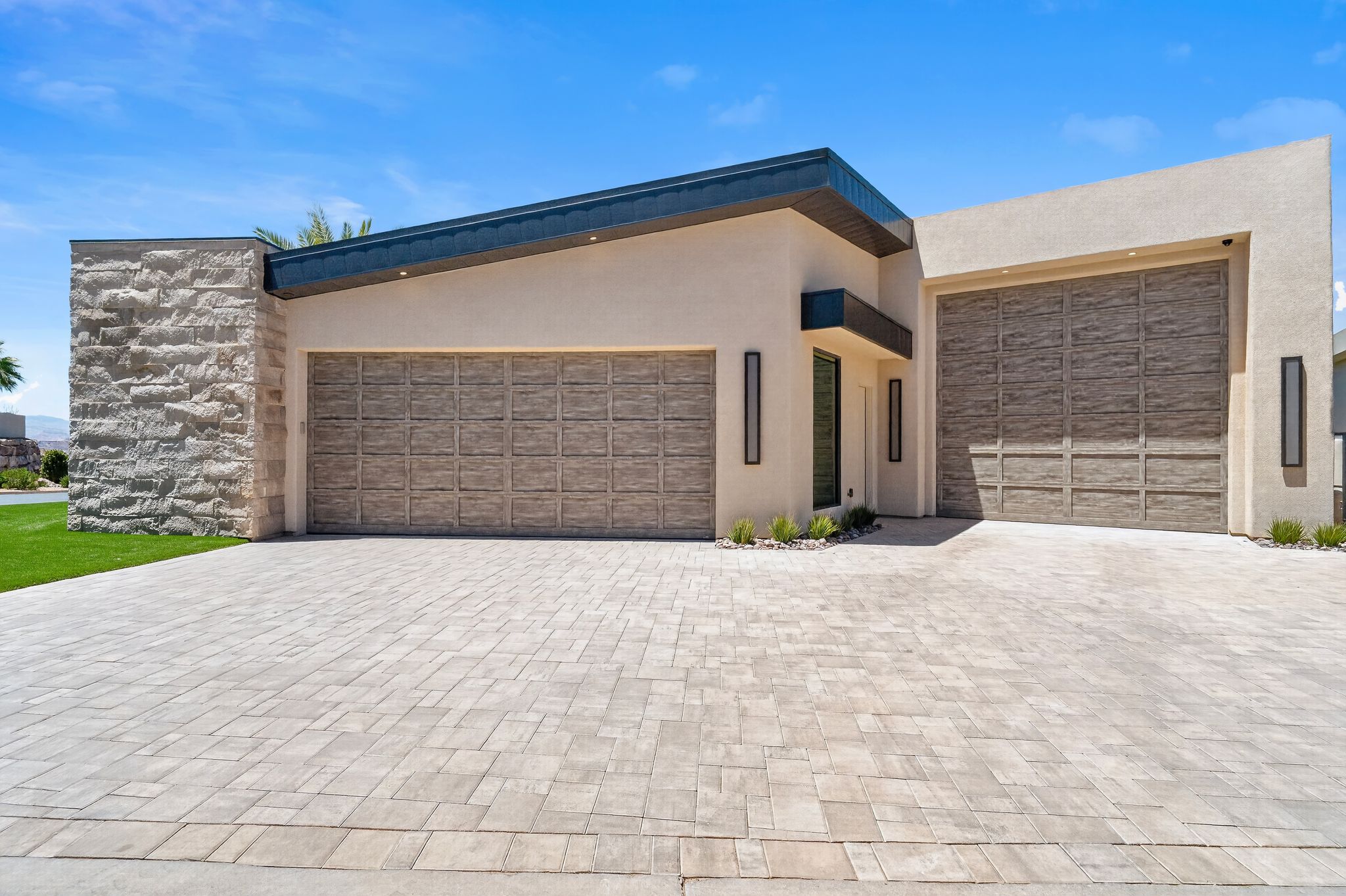 Modern exterior featuring a light-toned stucco finish, stacked stone accents, a multi-car paver driveway, sloped roofline, and integrated garage doors with a wood-finish pattern