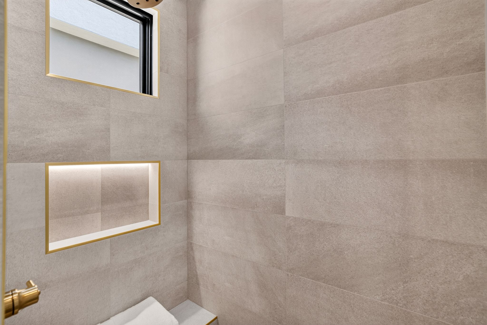 Tiled shower enclosure featuring a built-in bench, integrated niche with accent lighting, a window with golden trim, and a gold-toned shower fixture