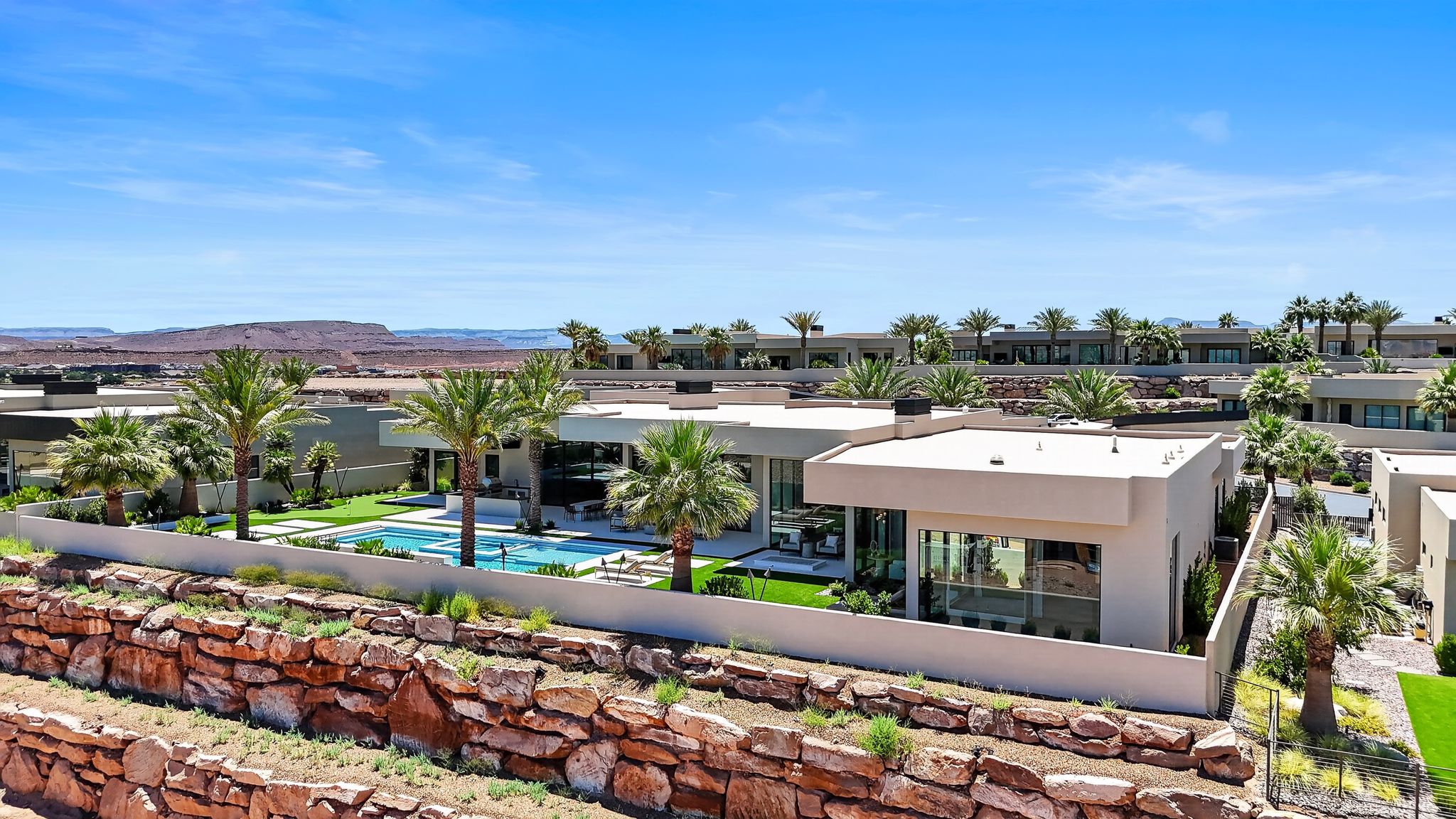 Modern architectural design featuring expansive glass panels, a private swimming pool, artificial turf landscaping, mature palm trees, and a terraced rock retaining wall