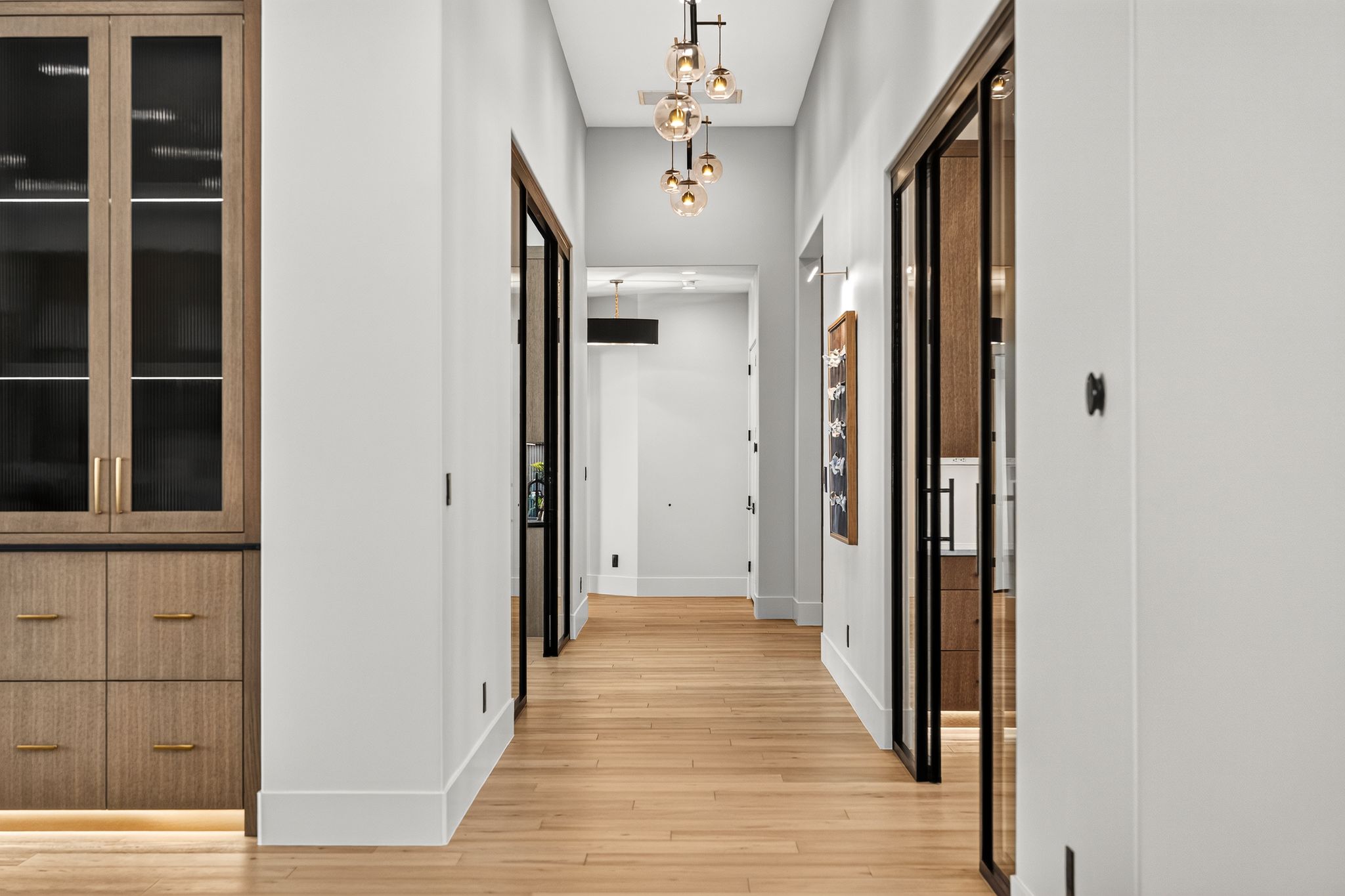 Hallway featuring wood-finish flooring, a contemporary multi-light pendant fixture, white walls, and dark-framed glass doors