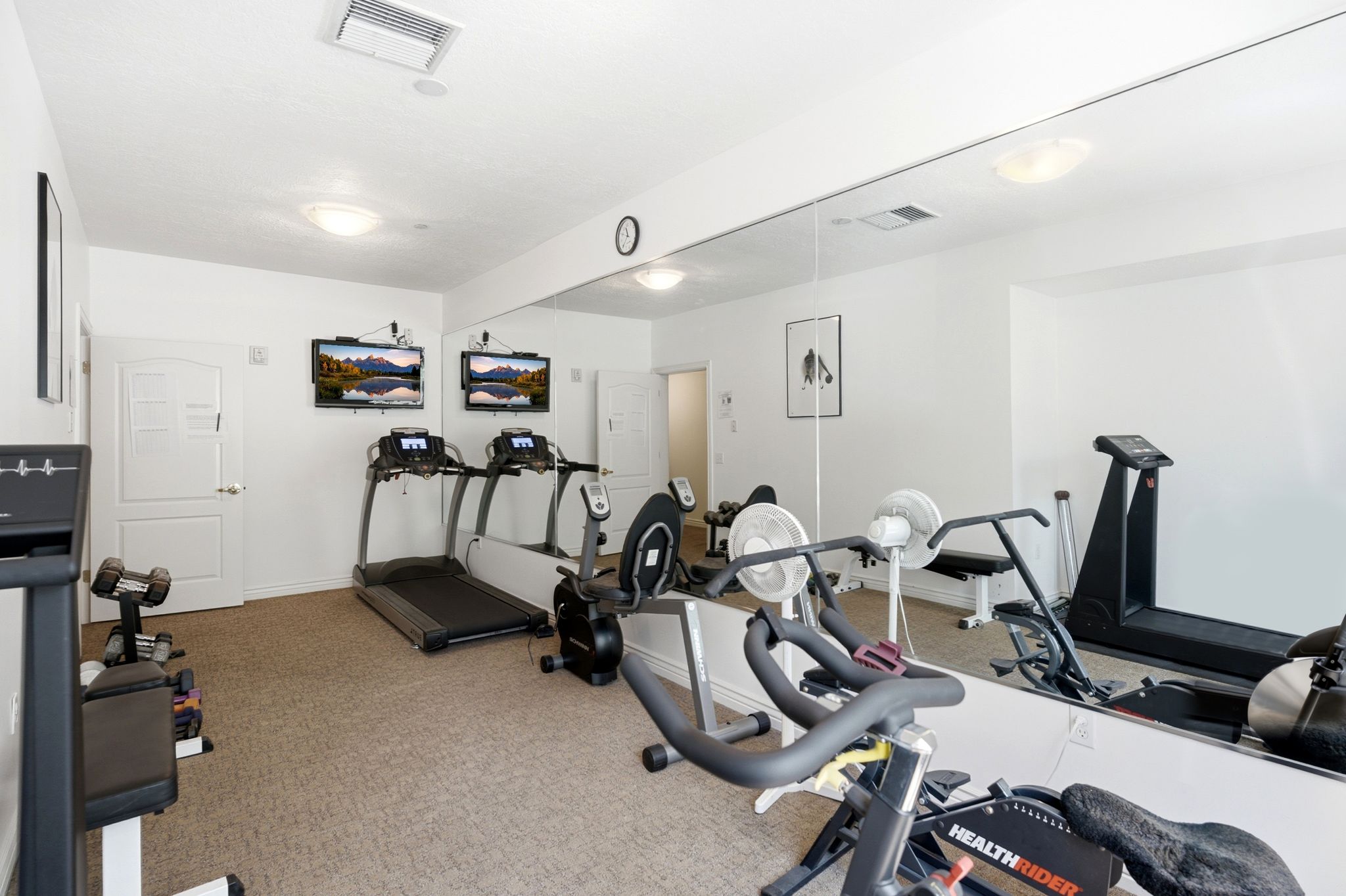 Dedicated fitness room featuring wall-mounted televisions, full-wall mirror, overhead lighting, and neutral carpeting