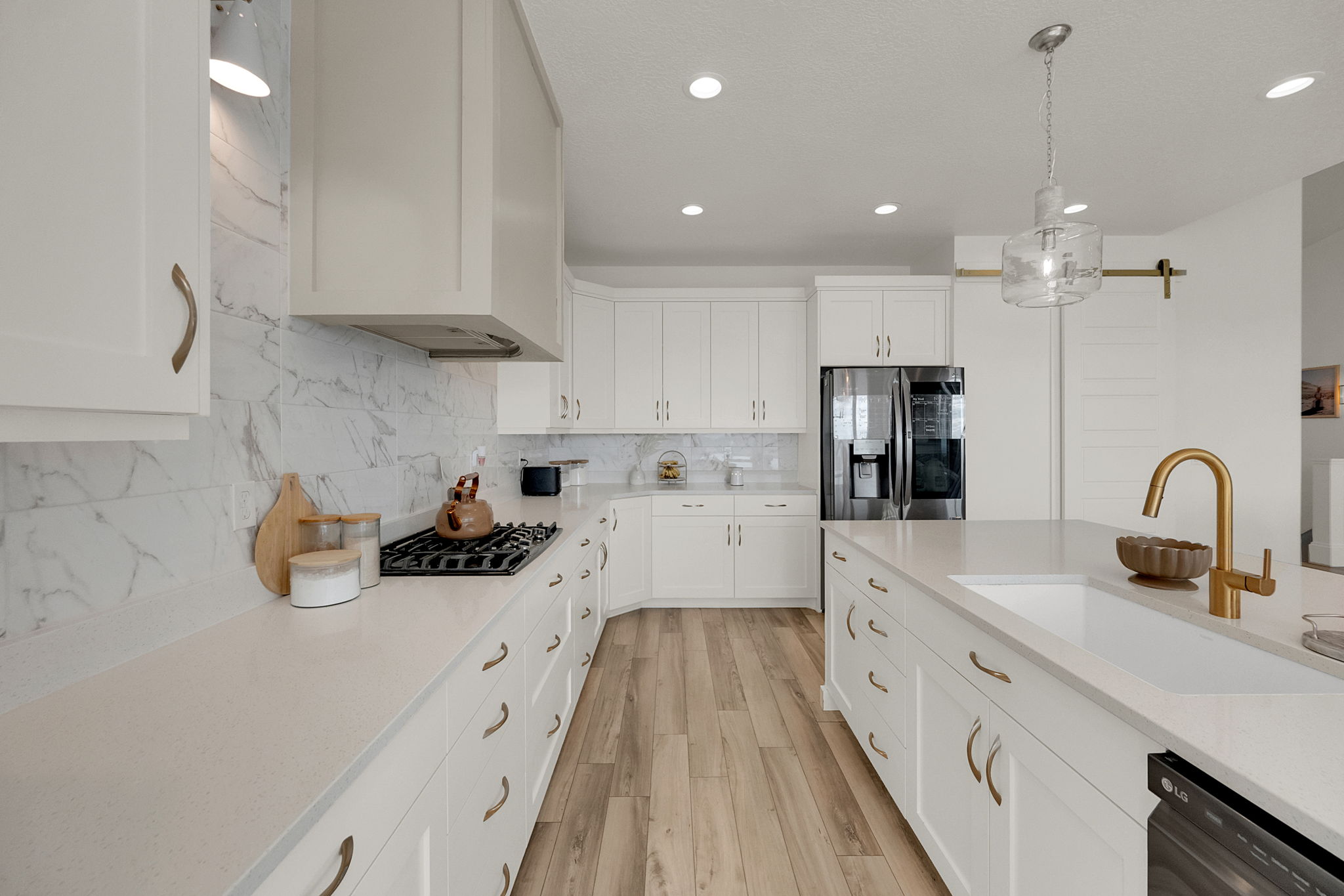 Contemporary kitchen featuring white cabinetry, light-toned countertops, a full height marble-look backsplash, wood-finish flooring, and a central island with an undermount sink and gold-toned faucet