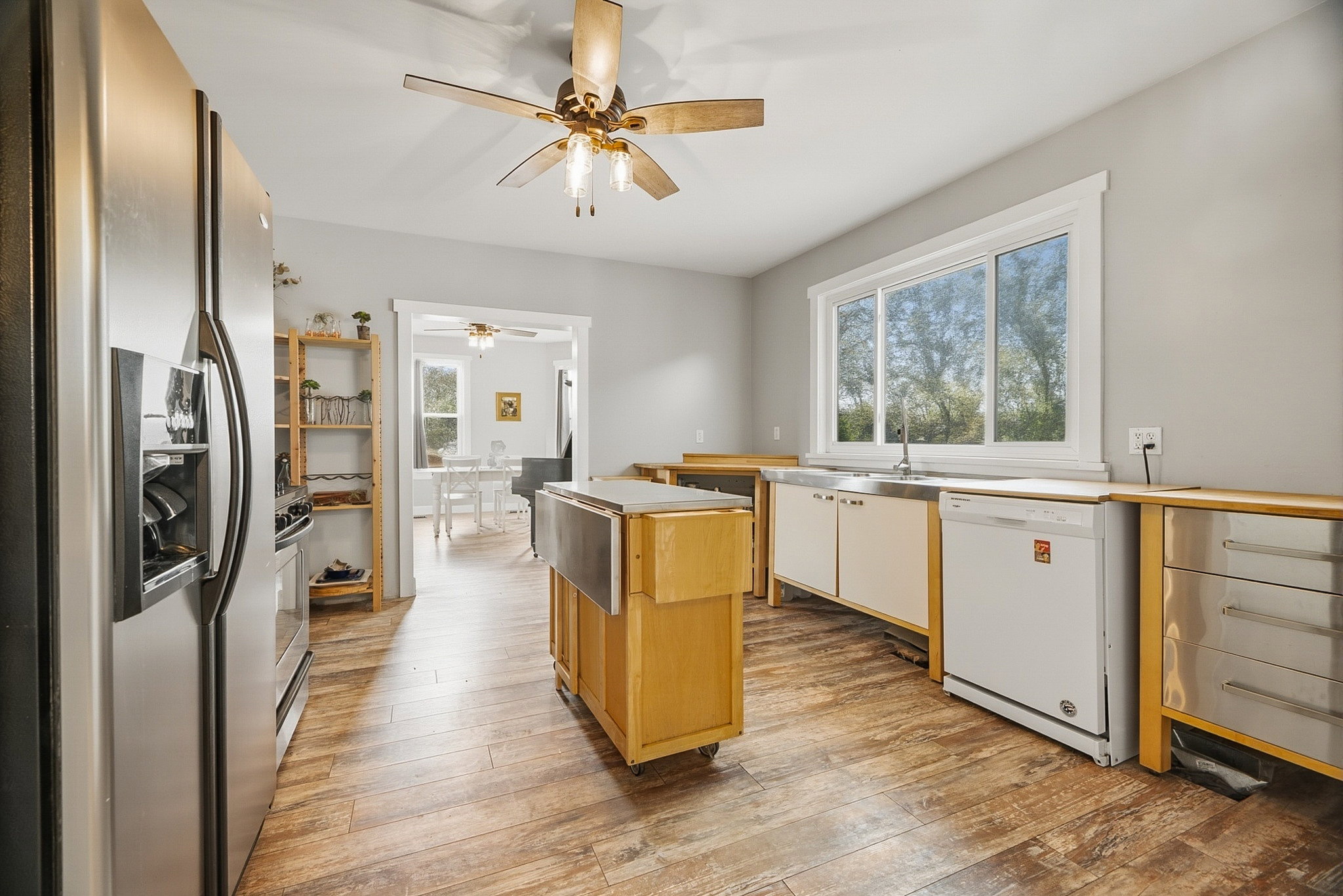 Open kitchen featuring wood-finish flooring, stainless steel refrigerator, white dishwasher, and a large window above the sink