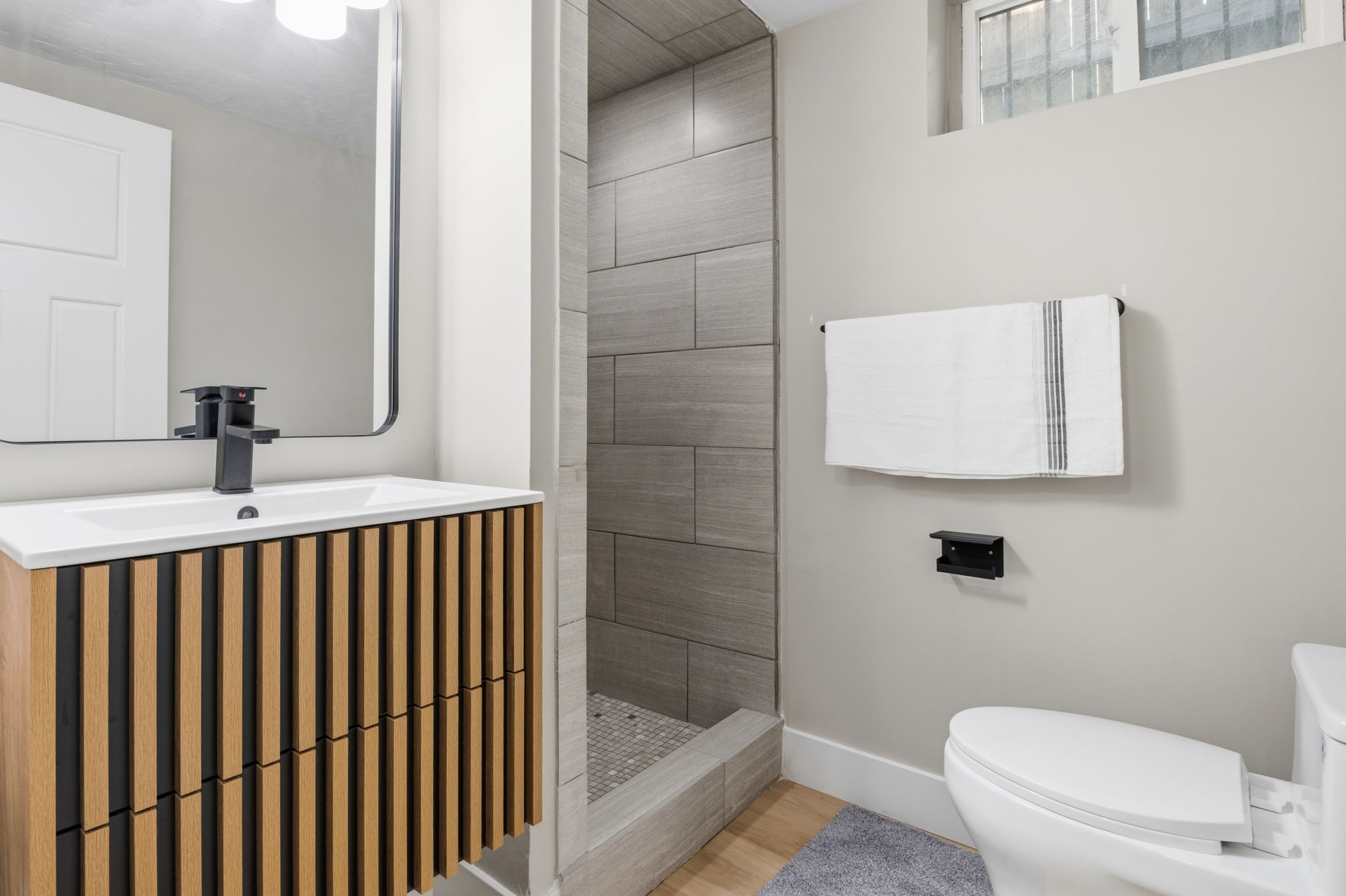 Basement  Second Modern bathroom featuring a wood-panel vanity with a white countertop and rectangular sink, matte black faucet, and a frameless mirror