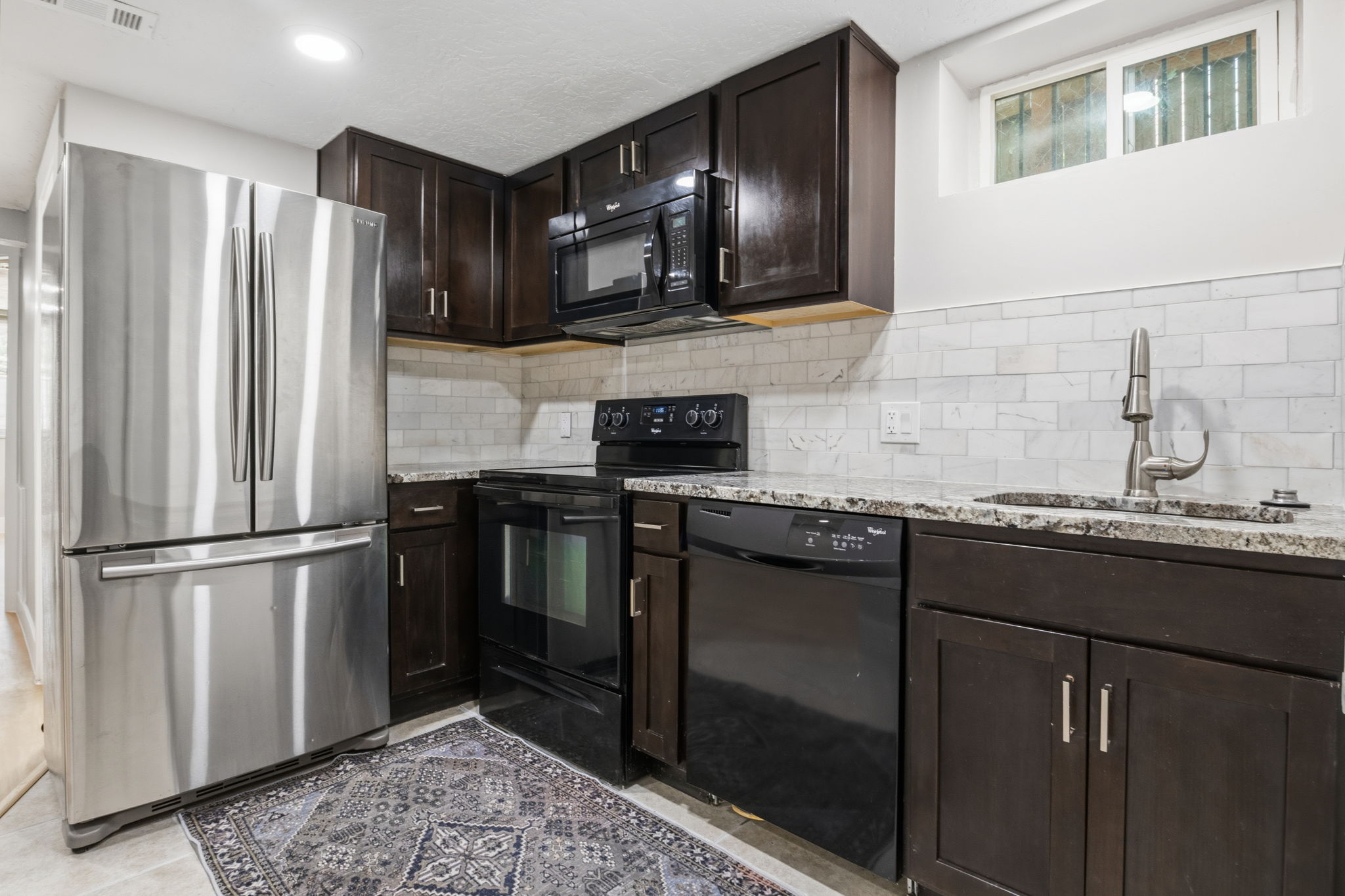 Basemet Kitchen featuring dark wood cabinetry, granite-finish countertops, stainless steel refrigerator, and subway tile backsplash