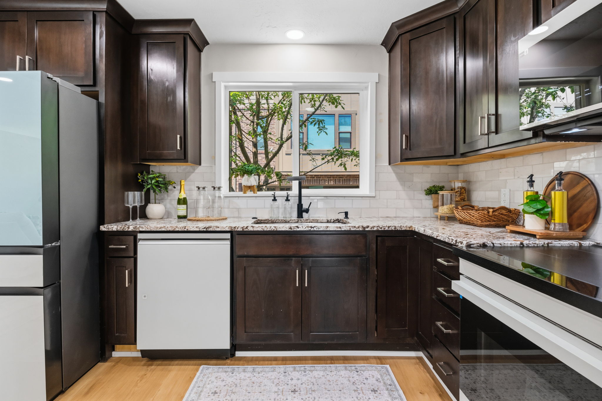 Kitchen featuring dark wood cabinetry, light-colored granite countertops, a white subway tile backsplash, a window above the sink, and wood-finish flooring