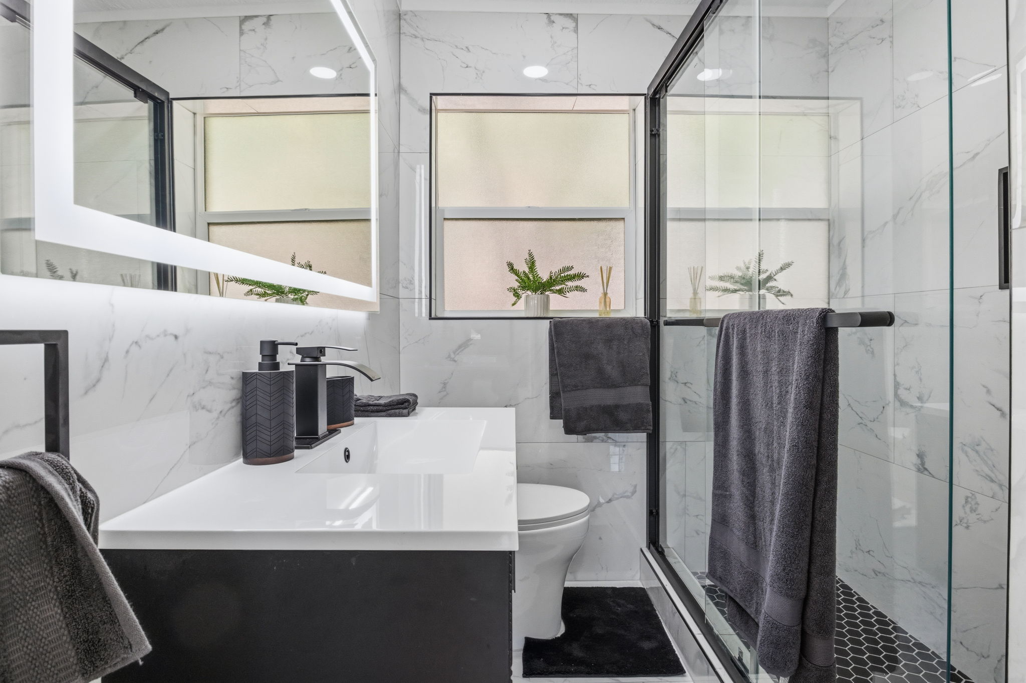 Contemporary bathroom featuring a black vanity with an integrated white sink, an illuminated framed mirror, and a glass-enclosed shower with black hexagonal tile flooring