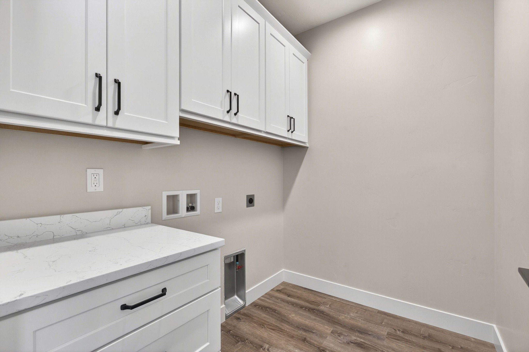 Laundry area featuring white shaker-style cabinetry with matte black hardware, a light-toned countertop with veining, and wood-finish flooring