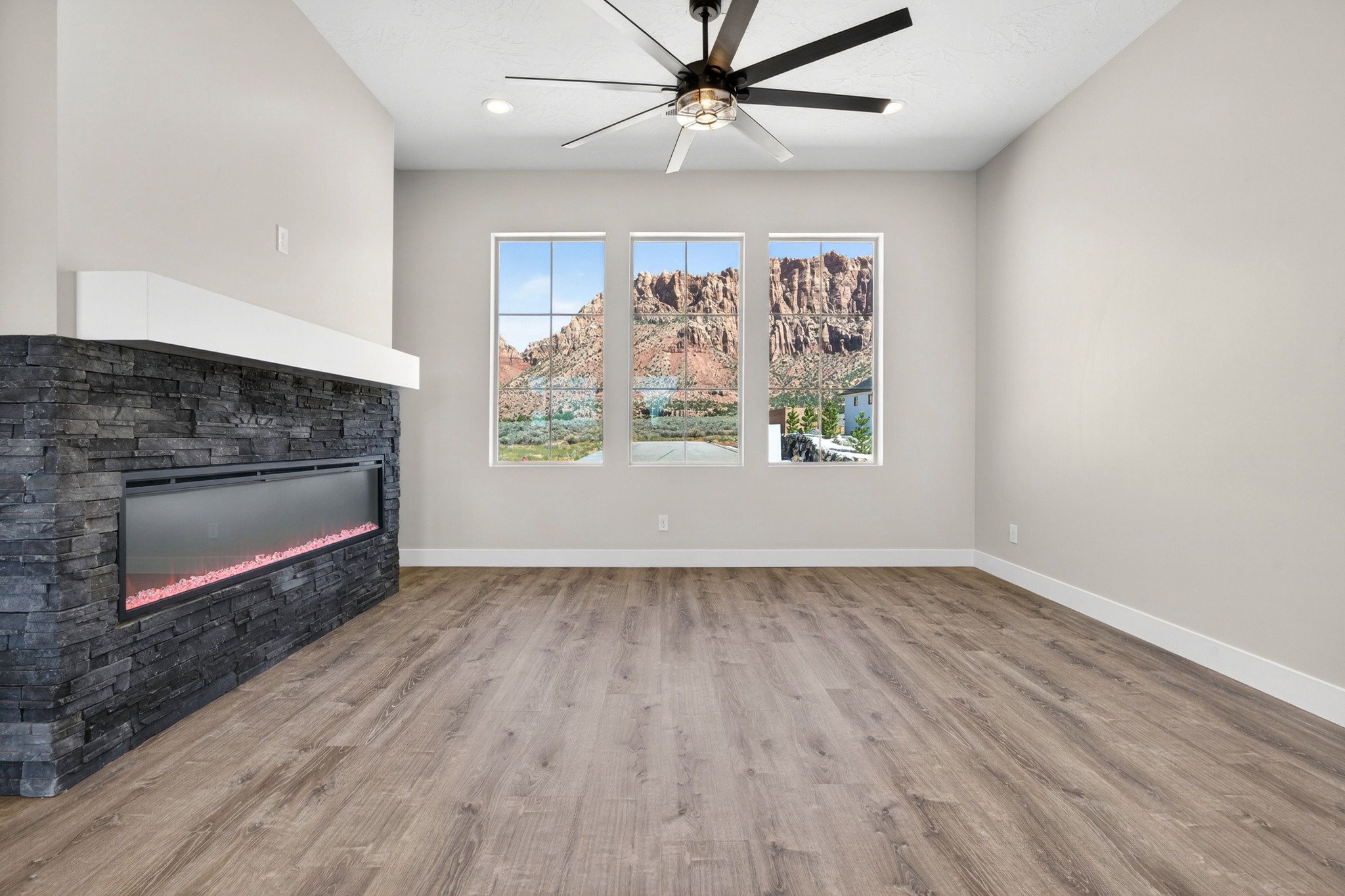 Spacious room featuring three large windows, a stone-clad linear fireplace with a white mantel, wood-finish flooring, and a modern ceiling fan