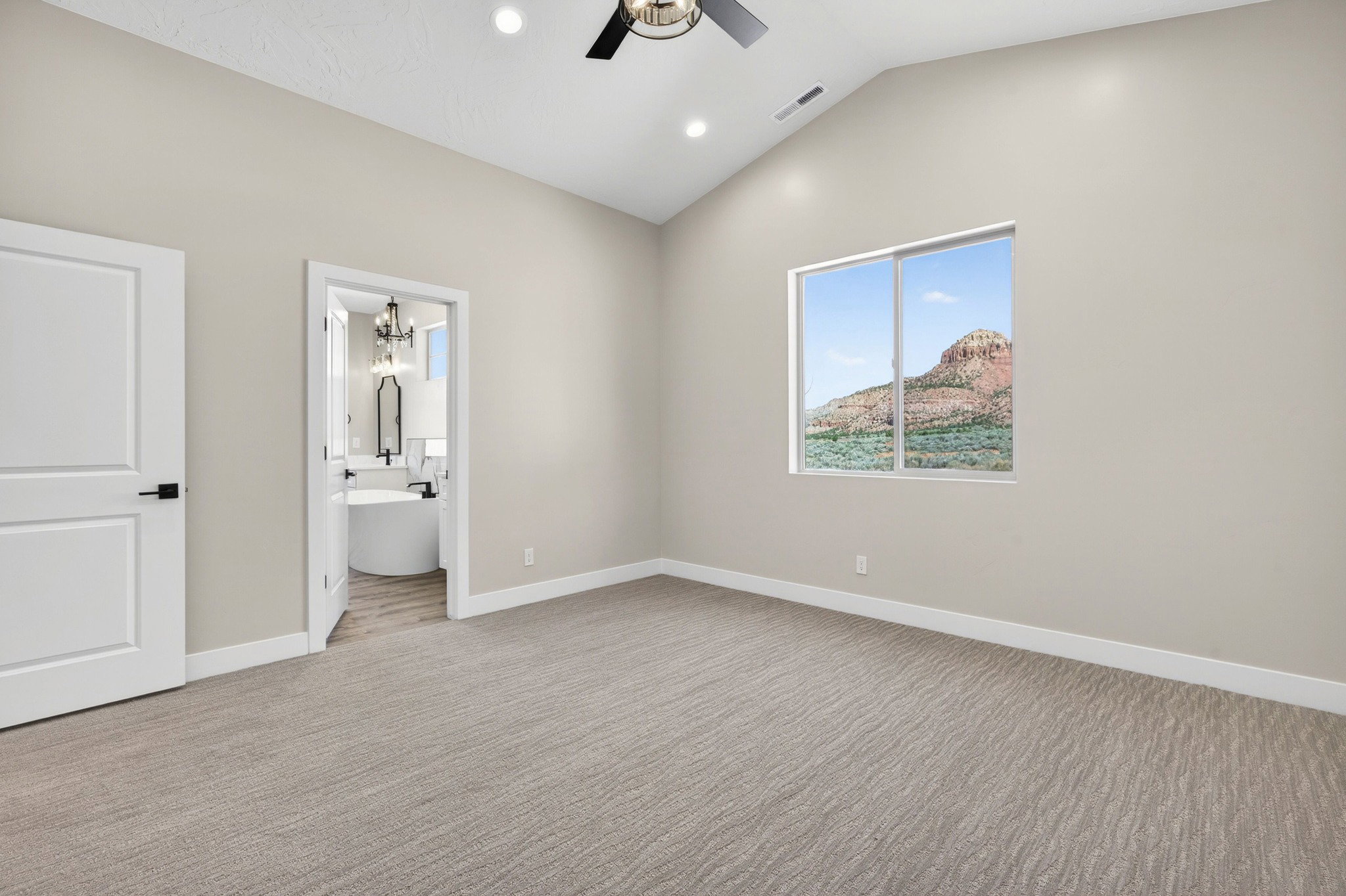 Vaulted ceiling bedroom with a patterned carpet, a large window showcasing mountain scenery, and an ensuite bathroom featuring a freestanding tub and chandelier