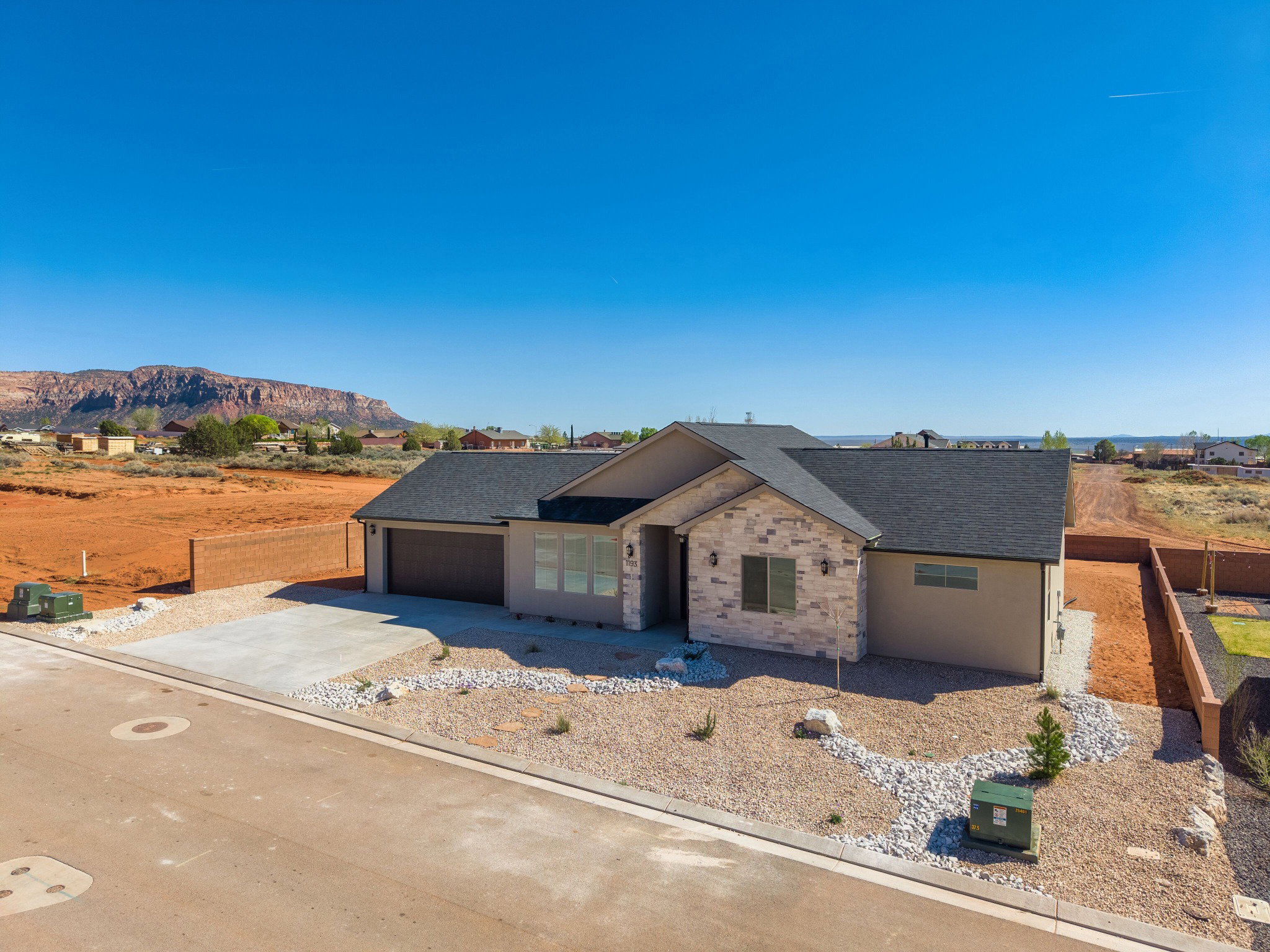 Modern ranch-style home featuring a two-car garage, stone and stucco exterior, dark shingle roof, and a gravel and rock landscaped front yard