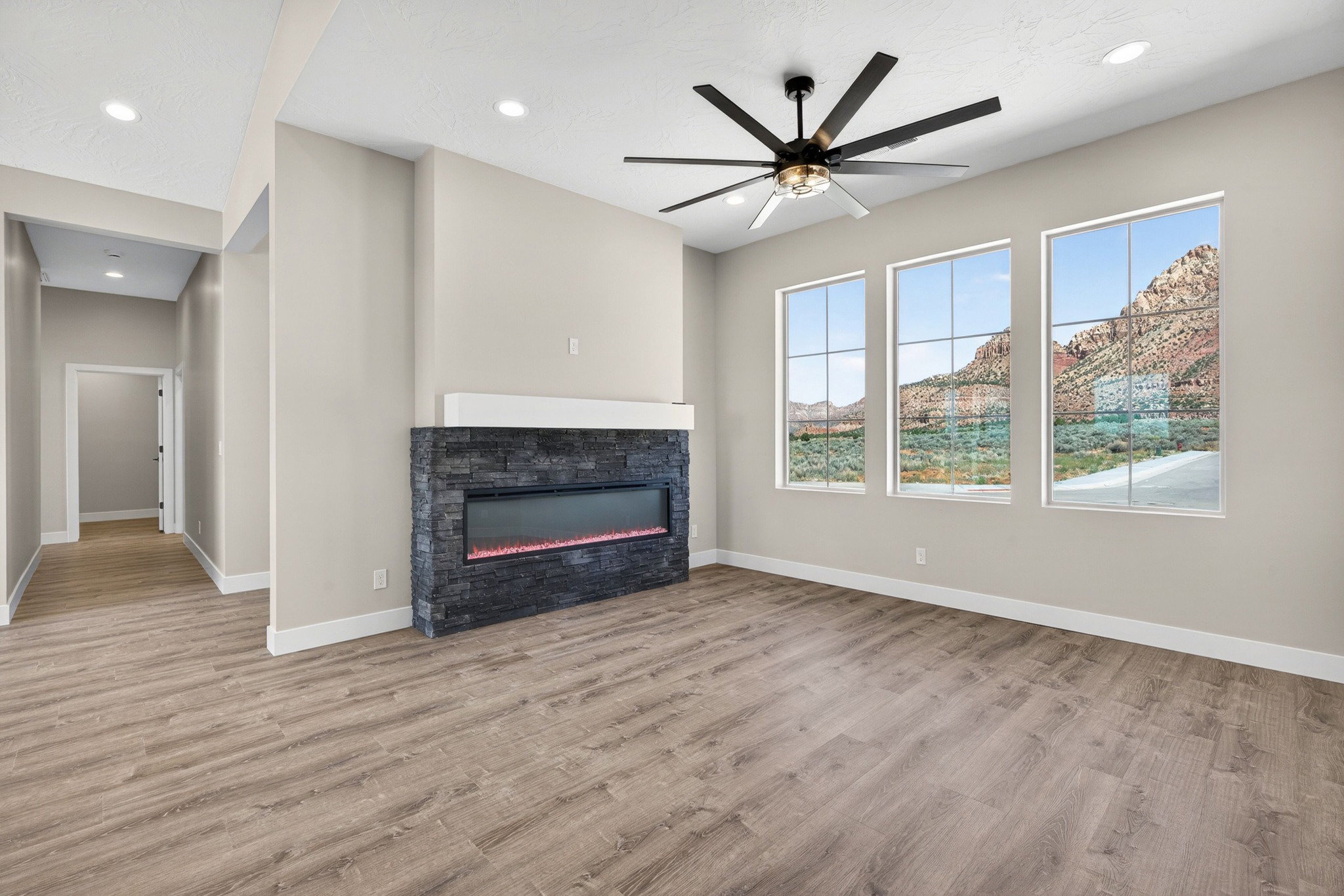 Spacious room featuring three large windows, a stone-clad linear fireplace, wood-finish flooring, and a contemporary ceiling fan