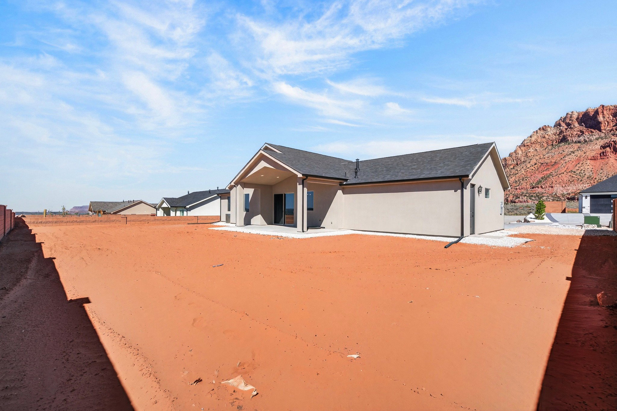 Spacious backyard with red earth, featuring a modern stucco exterior home with a covered patio, dark shingle roofing, and mountain backdrop